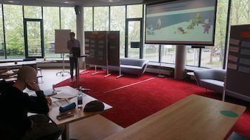 Moderator präsentiert in einem modernen, hell erleuchteten Raum mit großen Fenstern, einem digitalen Bildschirm, einem Whiteboard und Sitzgelegenheiten.