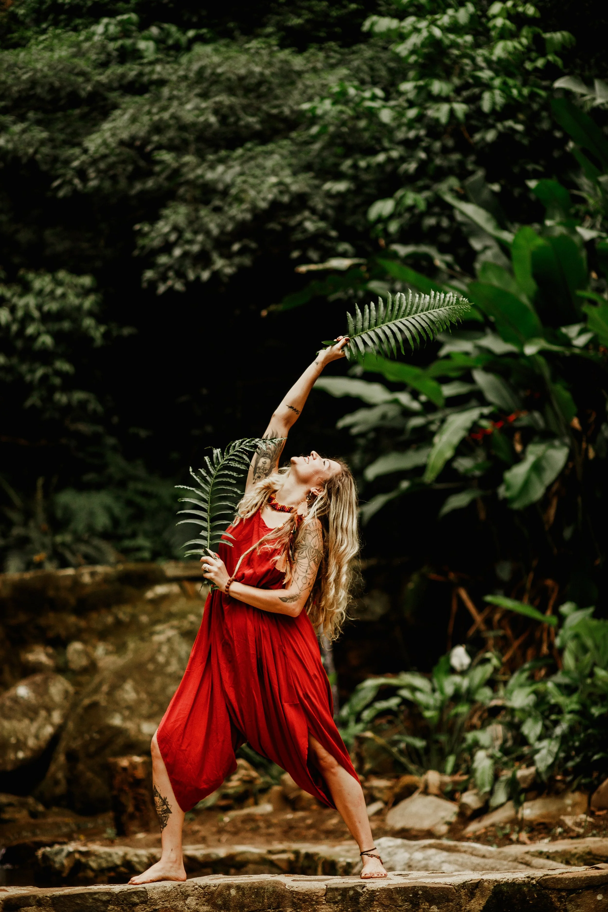 Mulher com cabelos longos loiros vestindo um vestido vermelho, de pé sobre uma pedra, segurando uma folha de samambaia com uma mão, em meio à natureza com vegetação densa ao fundo.