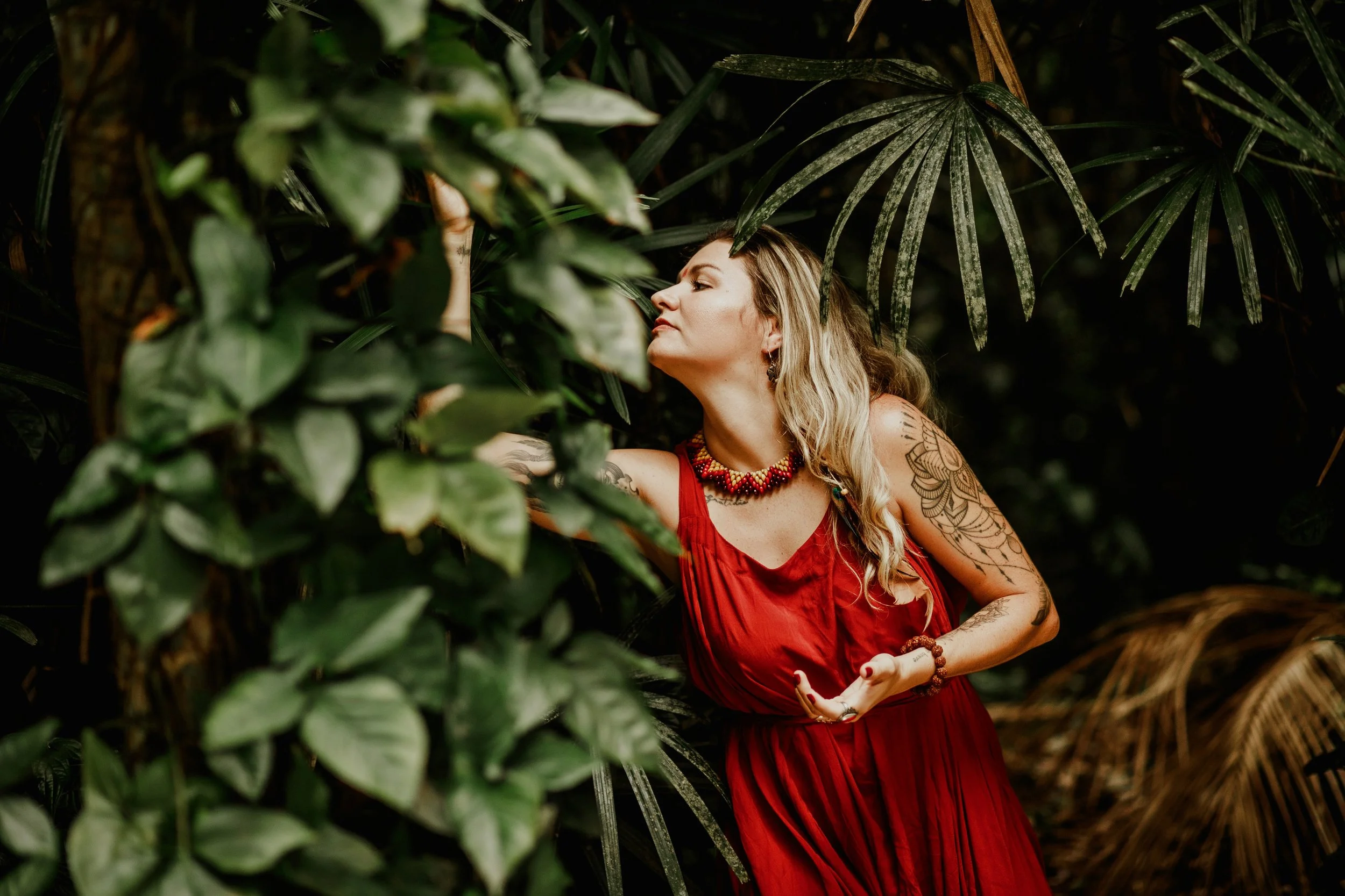 Mulher de cabelo longo e tatuagens na esquerda, vestindo vestido vermelho, entre folhas verdes em ambiente de floresta.