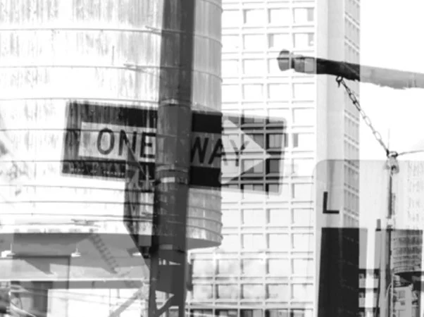 Black and white photo of a city street corner with a one-way street sign and a pole with a street name sign.