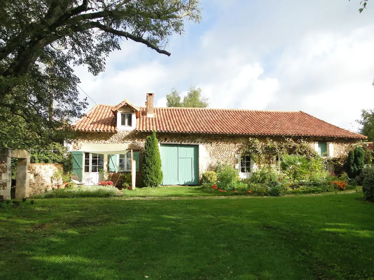Une maison de style rural avec des murs en pierre et un toit en tuiles rouges, entourée d’un jardin verdoyant et de plusieurs arbres et arbustes.