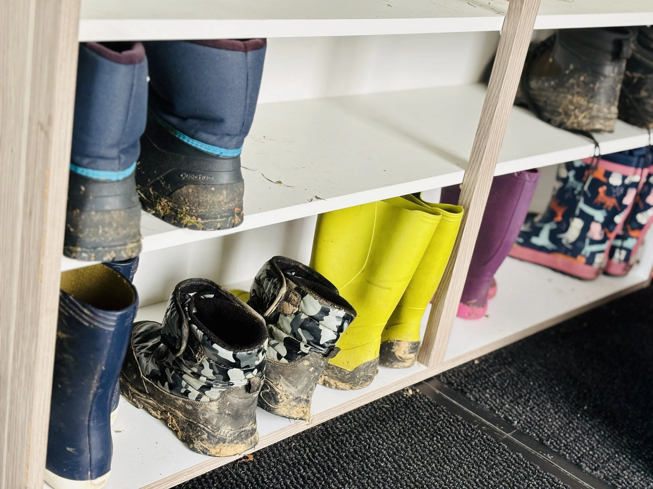 Chaussures de randonnée sales et bottes de pluie dans une étagère en bois, avec des chaussures de sport et un sac dans l'arrière-plan.