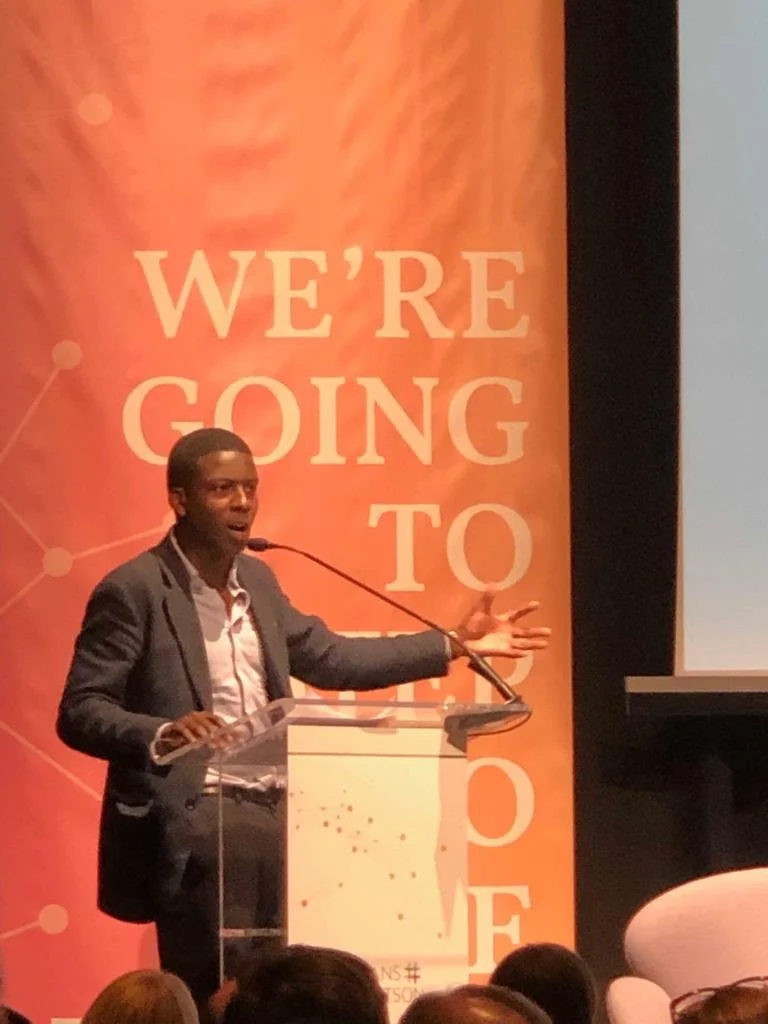 Man in a suit speaking at a podium with orange background that reads "We're Going To Do"
