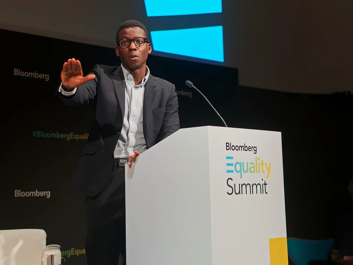 A man in a suit and glasses speaking at a podium with the words "Bloomberg Equality Summit" on it, gesturing with his hand. The background has a dark backdrop with the words "Bloomberg" and a large blue logo.