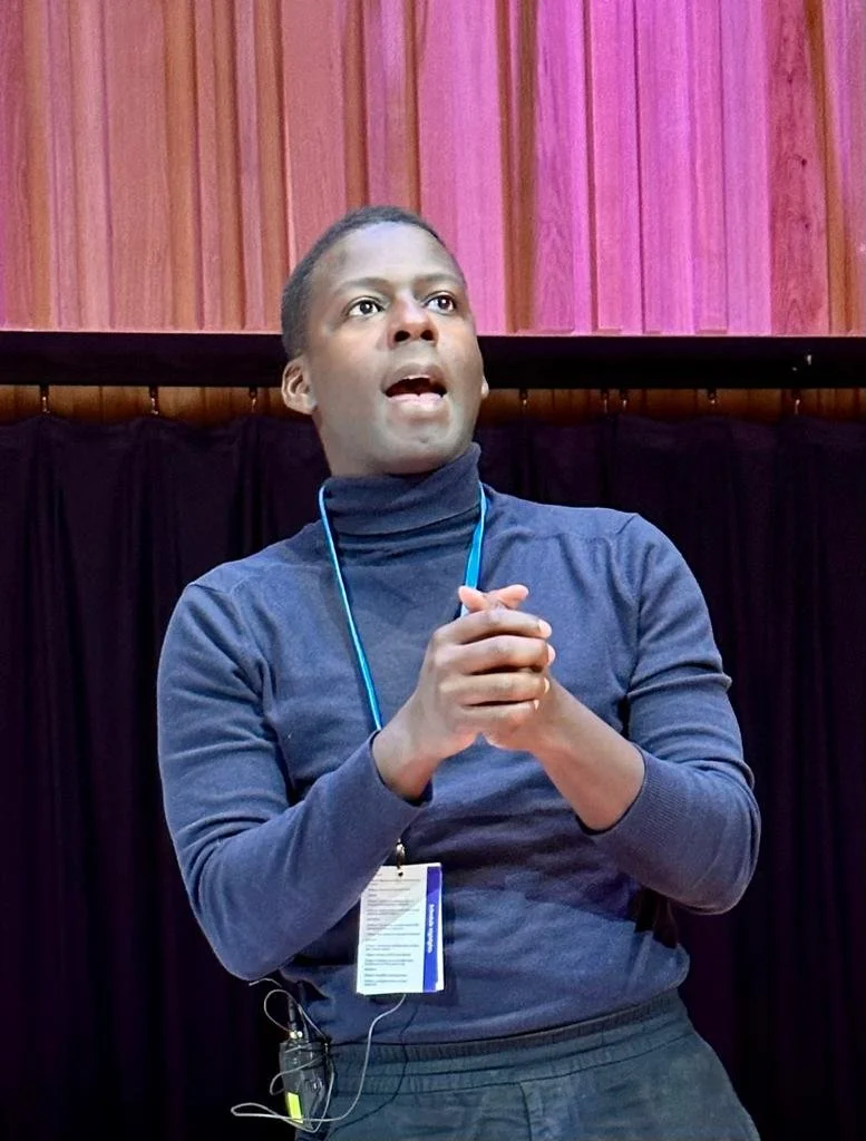 A person speaking or presenting indoors, wearing a navy blue turtleneck and a blue lanyard with an ID badge, standing in front of a black curtain with wooden paneling above.