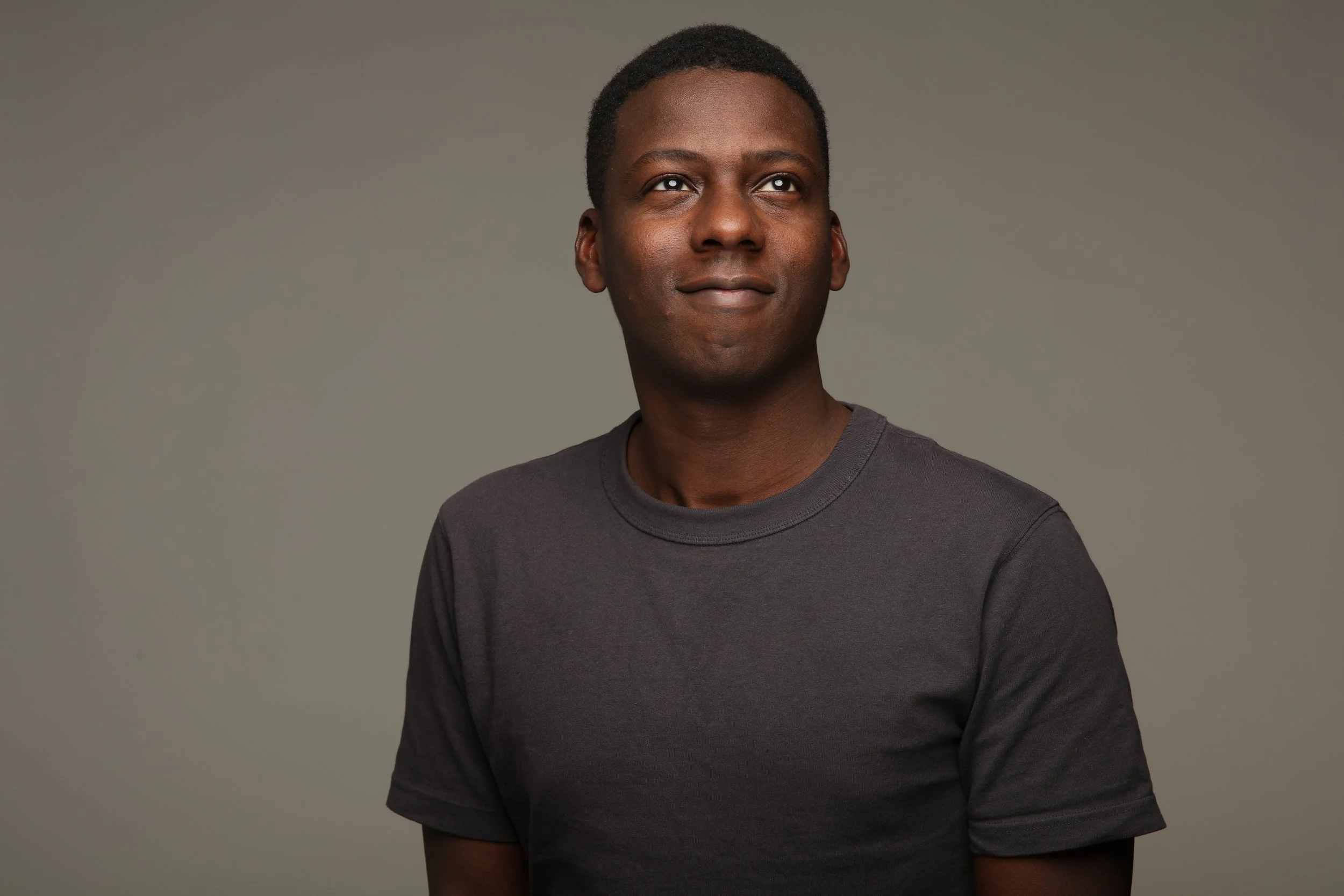 Portrait of a young African American man with short black hair, wearing a dark gray t-shirt, looking to his right against a neutral gray background.