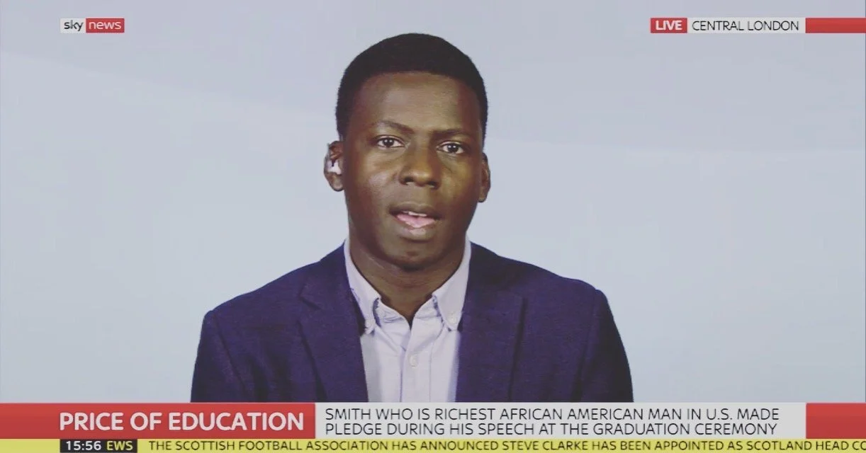 A young Black man in a navy blazer and white shirt speaking on a news broadcast from Central London, with a headlines about the price of education and a caption about Smith, the richest African American man in the U.S., at the graduation ceremony.