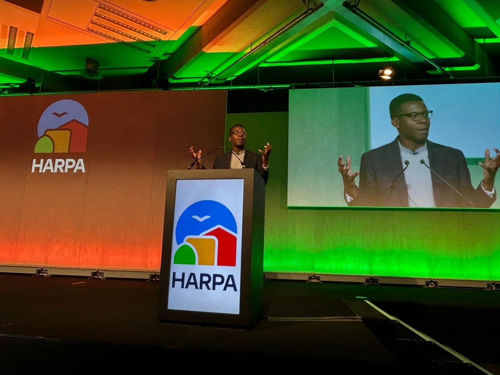 A man in a dark blazer and glasses is giving a speech at aHARPA conference, gesturing with his hands. There is a large screen behind him showing his image, and a podium with the HARPA logo, which depicts a stylized illustration of houses, a bird, and a sun or moon, with the word HARPA underneath. The background and lighting of the stage involve green and orange colors.