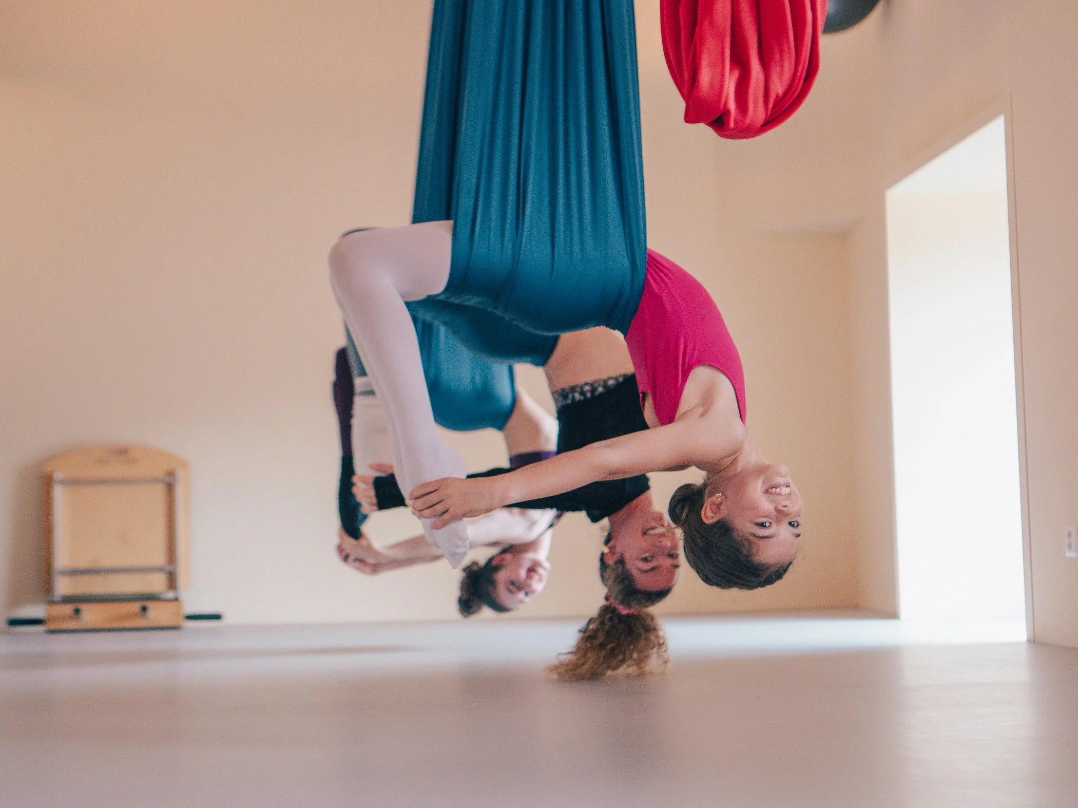 A group of people practicing aerial yoga, hanging upside down with their legs wrapped in fabric and holding onto each other's hands in a yoga studio.