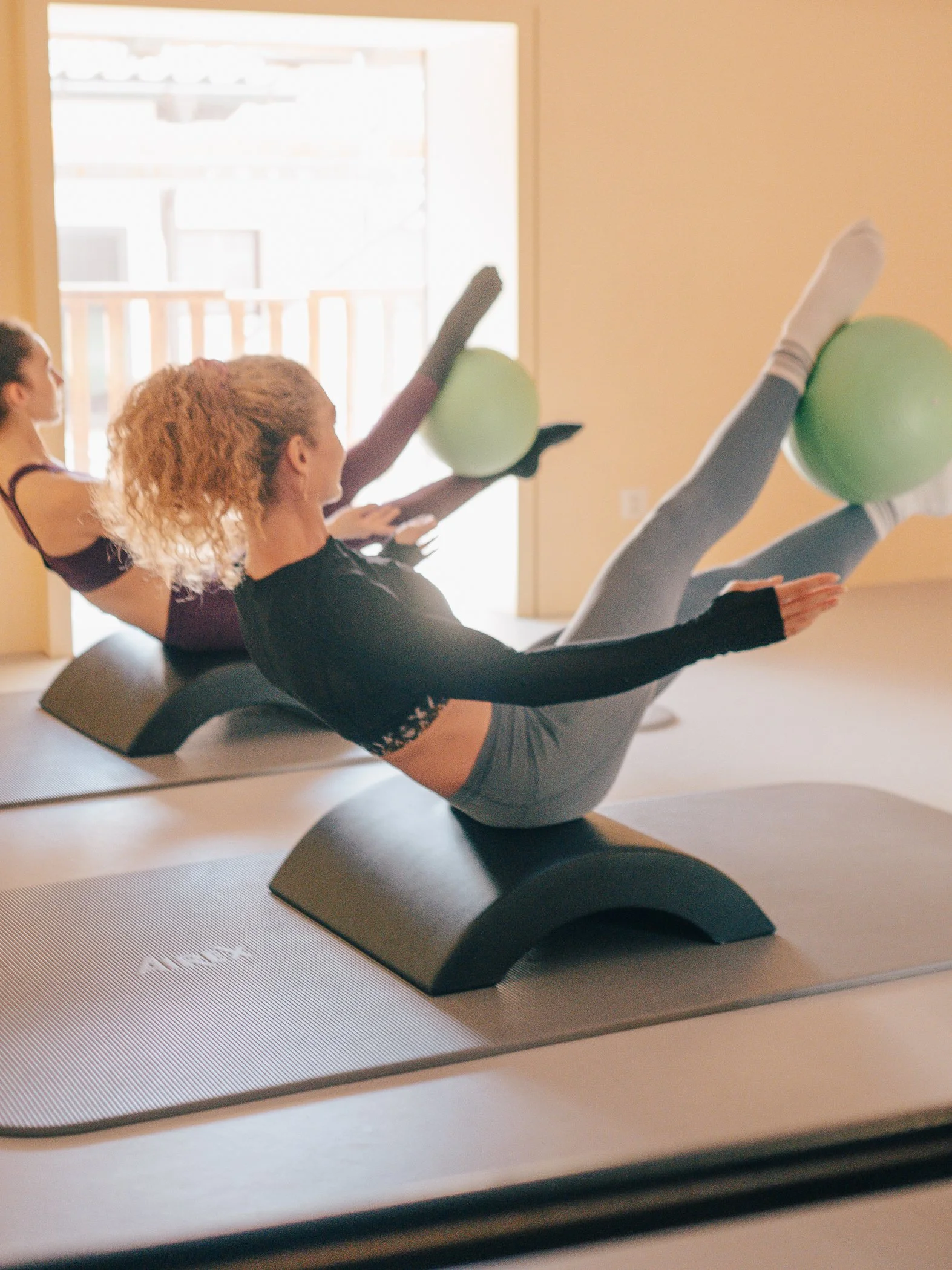 Three women practicing yoga on mats, lifting legs with stability balls, in a bright room with a sliding door leading outside.