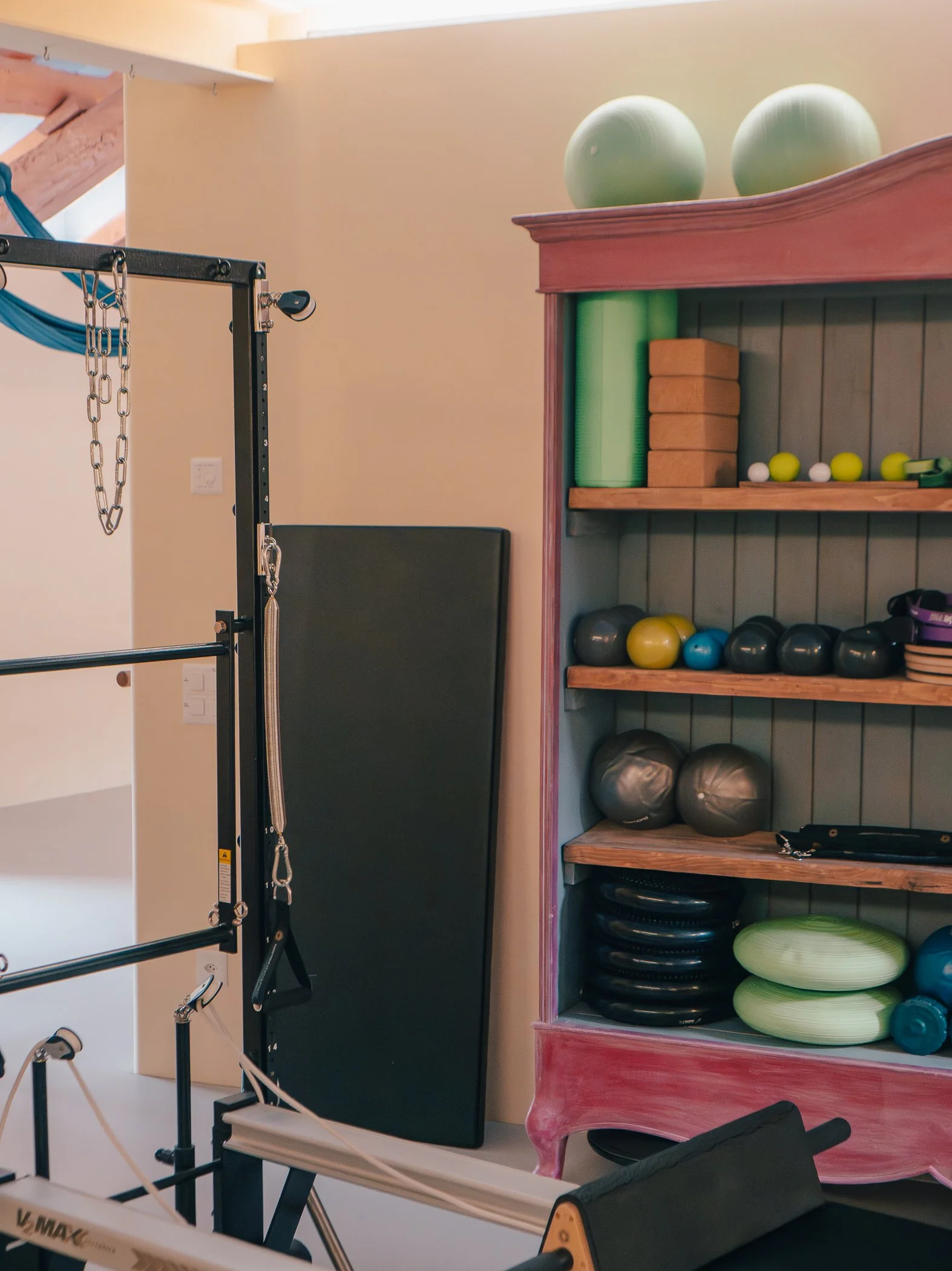 A gym corner with workout equipment including foam rollers, stability balls, kettlebells, and a wooden shelf, with part of a Pilates reformer machine visible in the foreground.