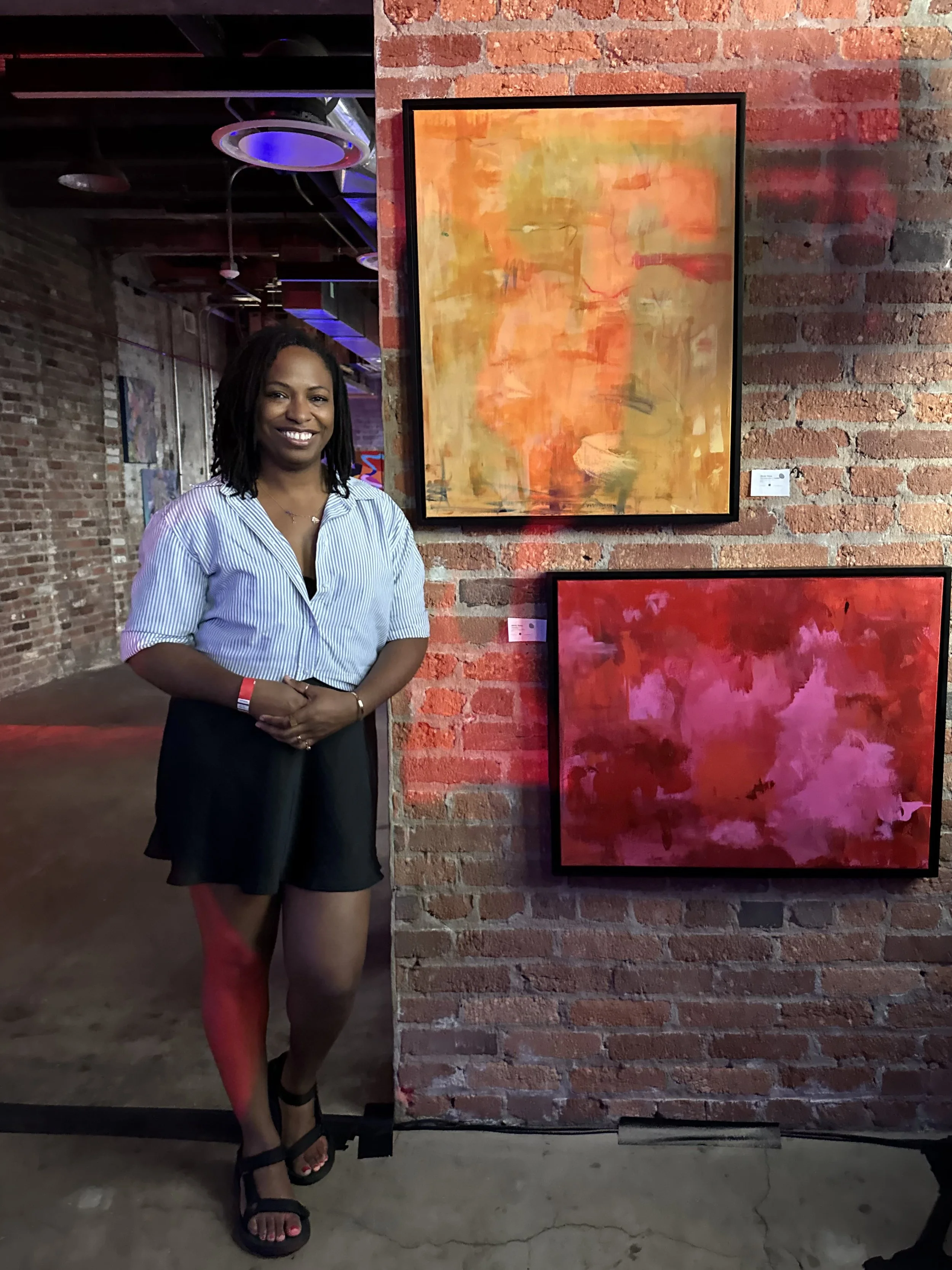 A woman with dark locs, wearing a light blue striped shirt and black skirt, standing next to a wall with red brick and abstract paintings in orange, red, and pink colors in an art gallery or exhibition space.