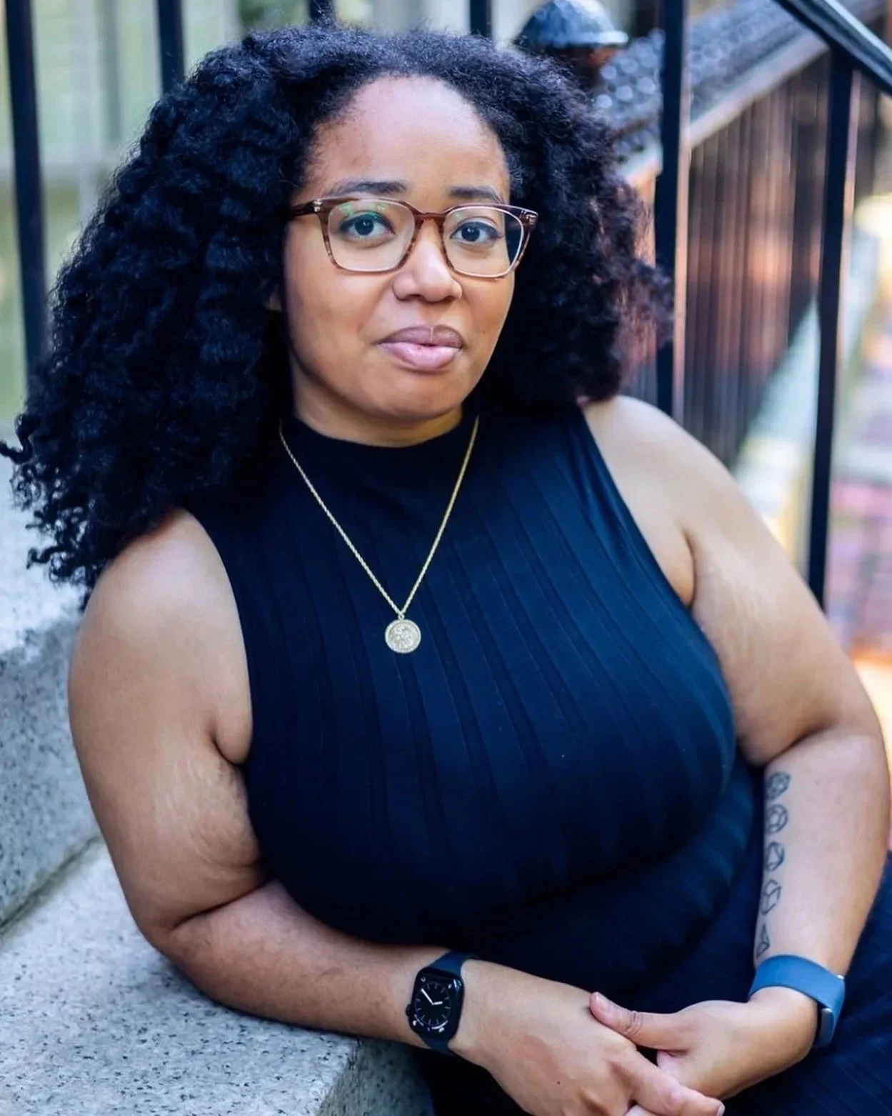 A woman with curly black hair, glasses, and a black sleeveless top standing outdoors near a railing and a concrete surface.