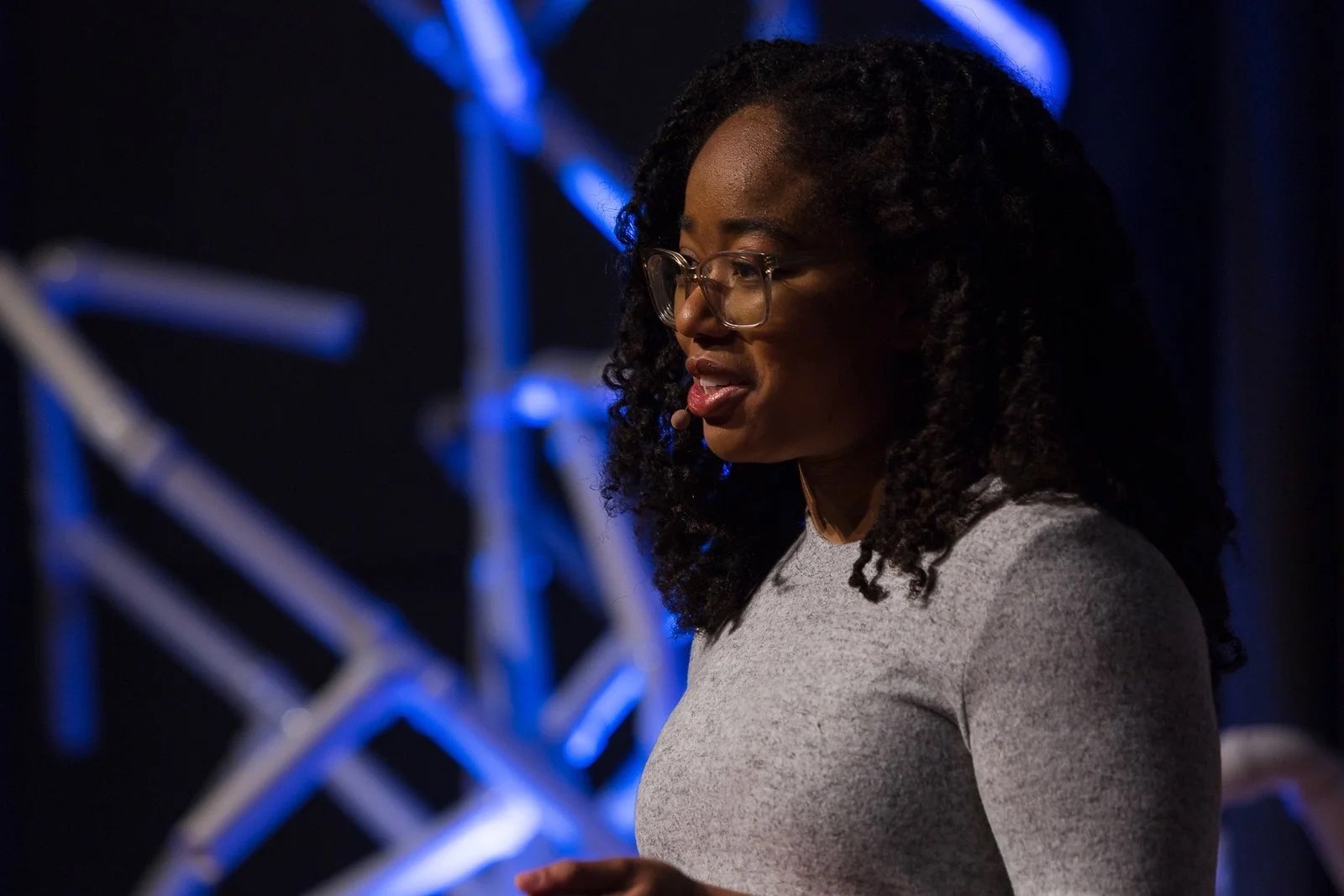 A woman with glasses and curly hair, wearing a gray shirt, appears to be speaking or presenting in a dark space with blue stage lighting and metal structures in the background.