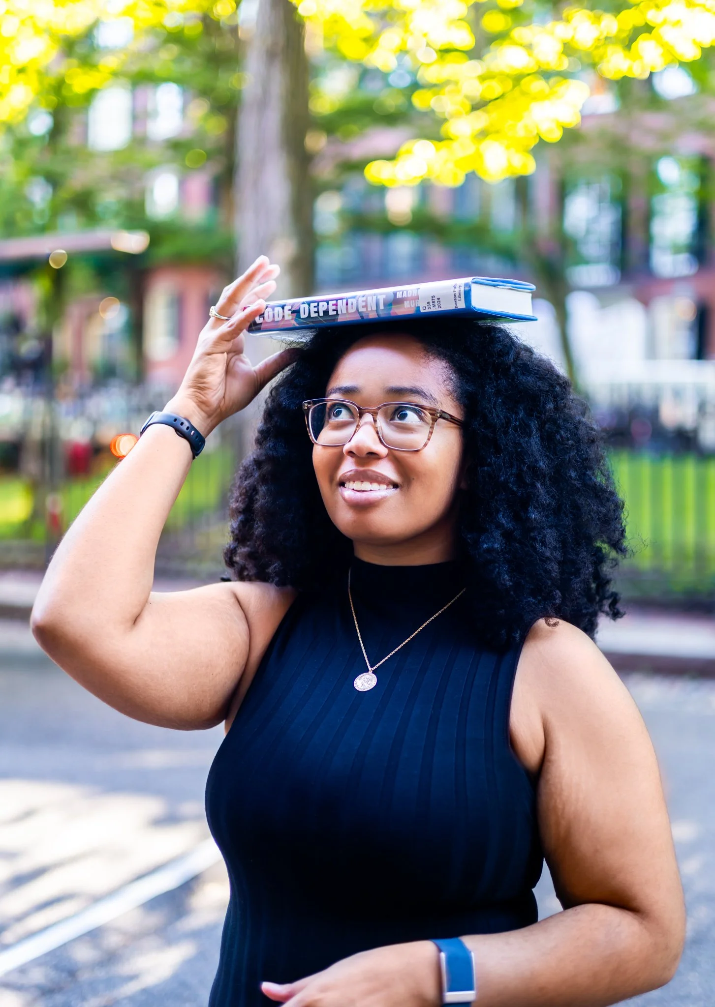 Young woman with curly black hair and glasses, wearing a black sleeveless top and a necklace, balancing a book titled 'Code Dependent' on her head outdoors during daytime.