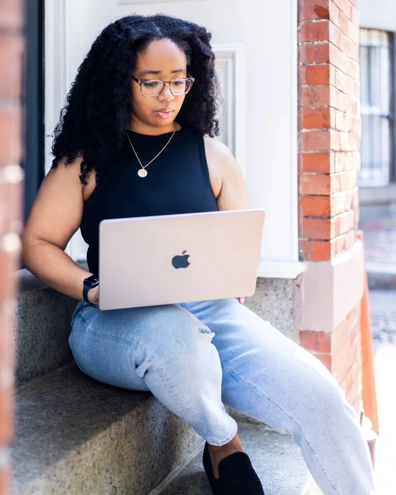 A young woman with curly hair, glasses, and a black sleeveless top sitting on concrete steps working on a silver MacBook laptop.