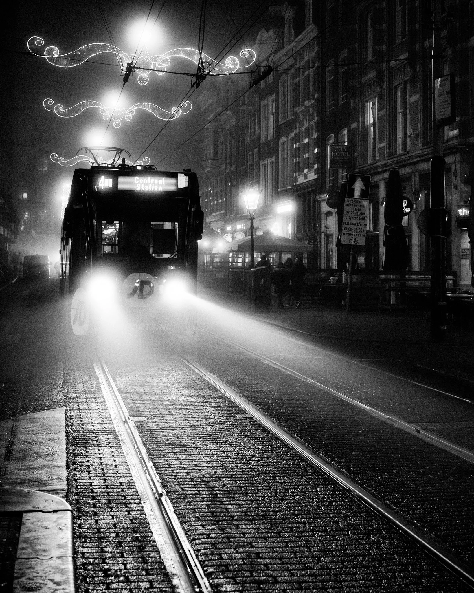 Black-and-white nighttime photo of a tram traveling on wet city tracks beneath festive lights, with historic buildings and a hotel sign in the background.
