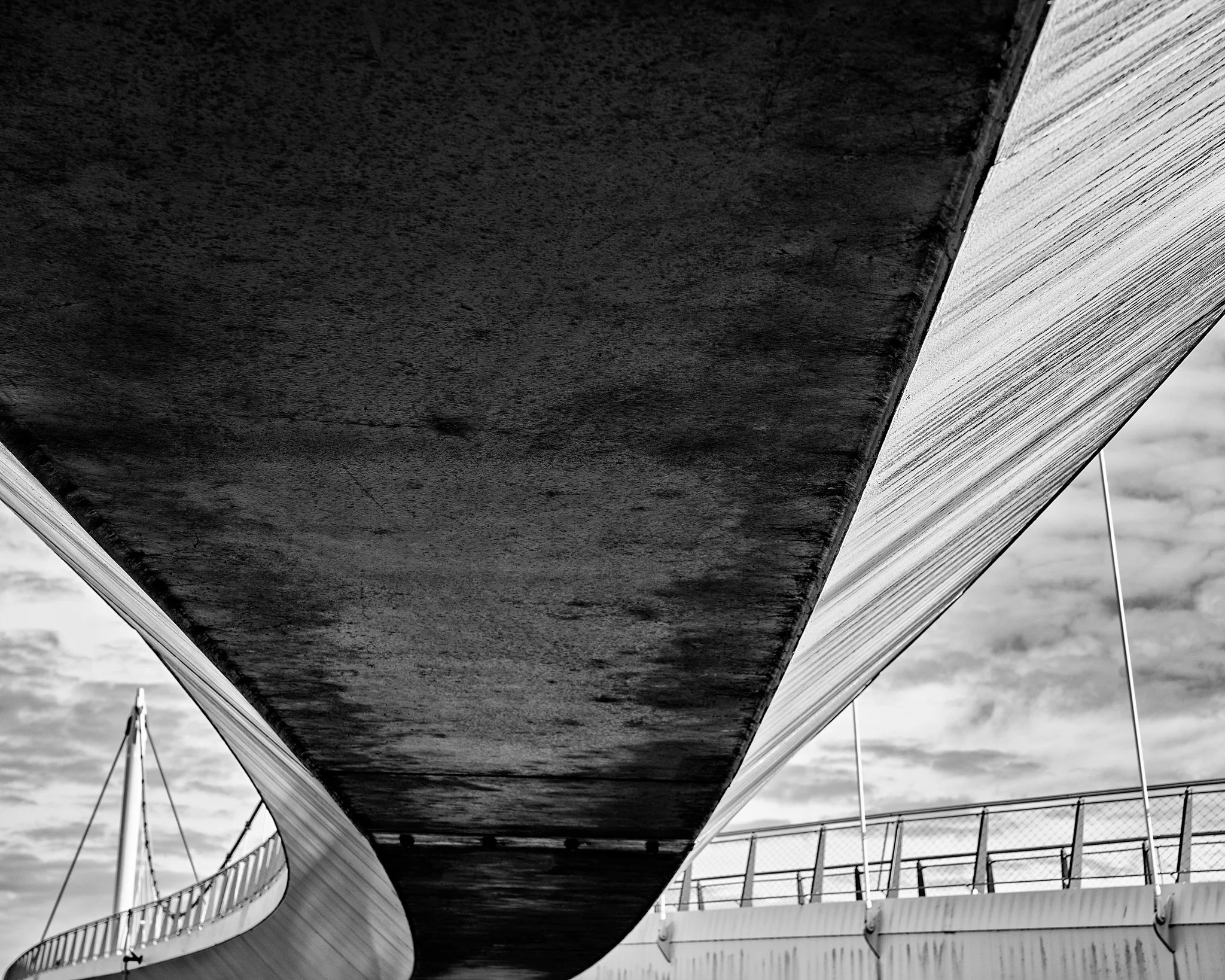 Black-and-white low-angle view beneath a large modern bridge with curved supports, railings, cables, and cloudy sky.