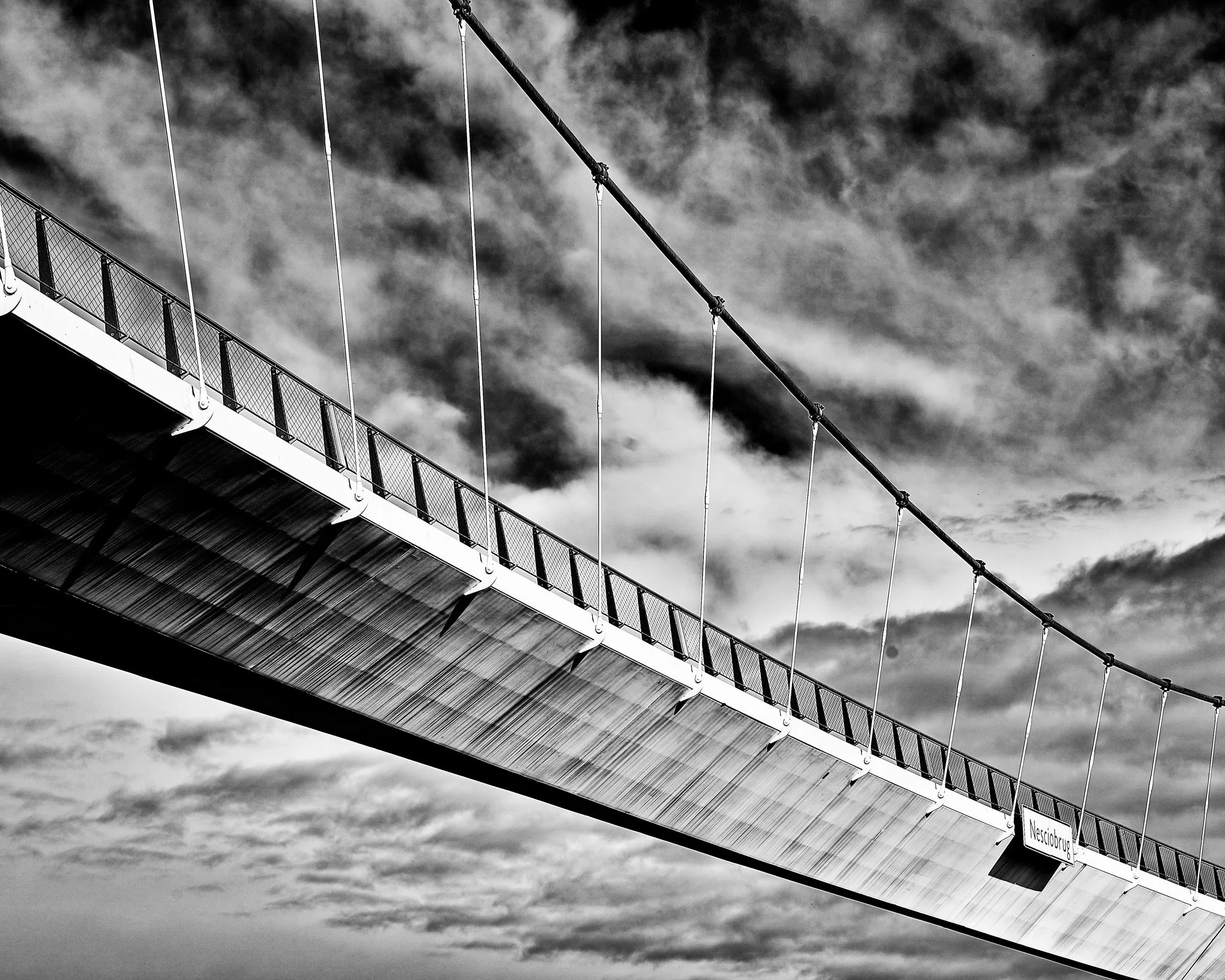 Black-and-white view of a suspension bridge deck and cables against a cloudy sky
