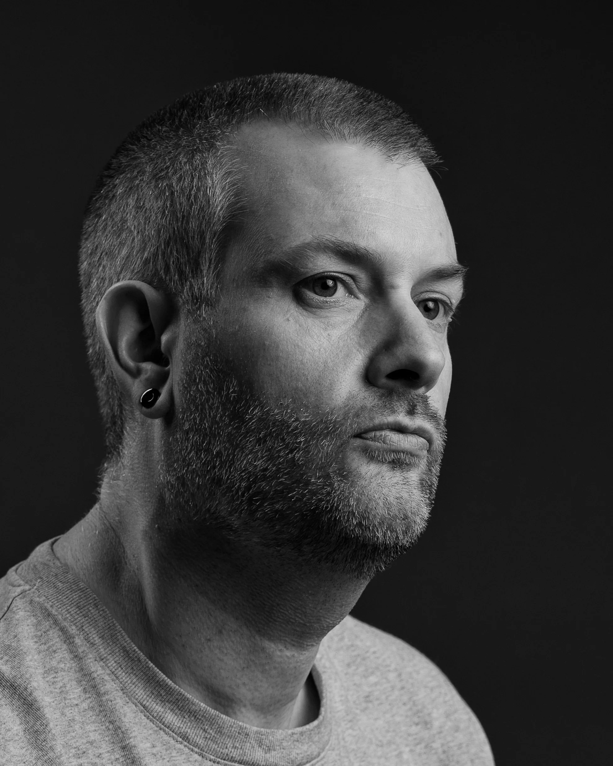 Black-and-white close-up portrait of a bearded man with short hair and a stretched earlobe plug against a dark background