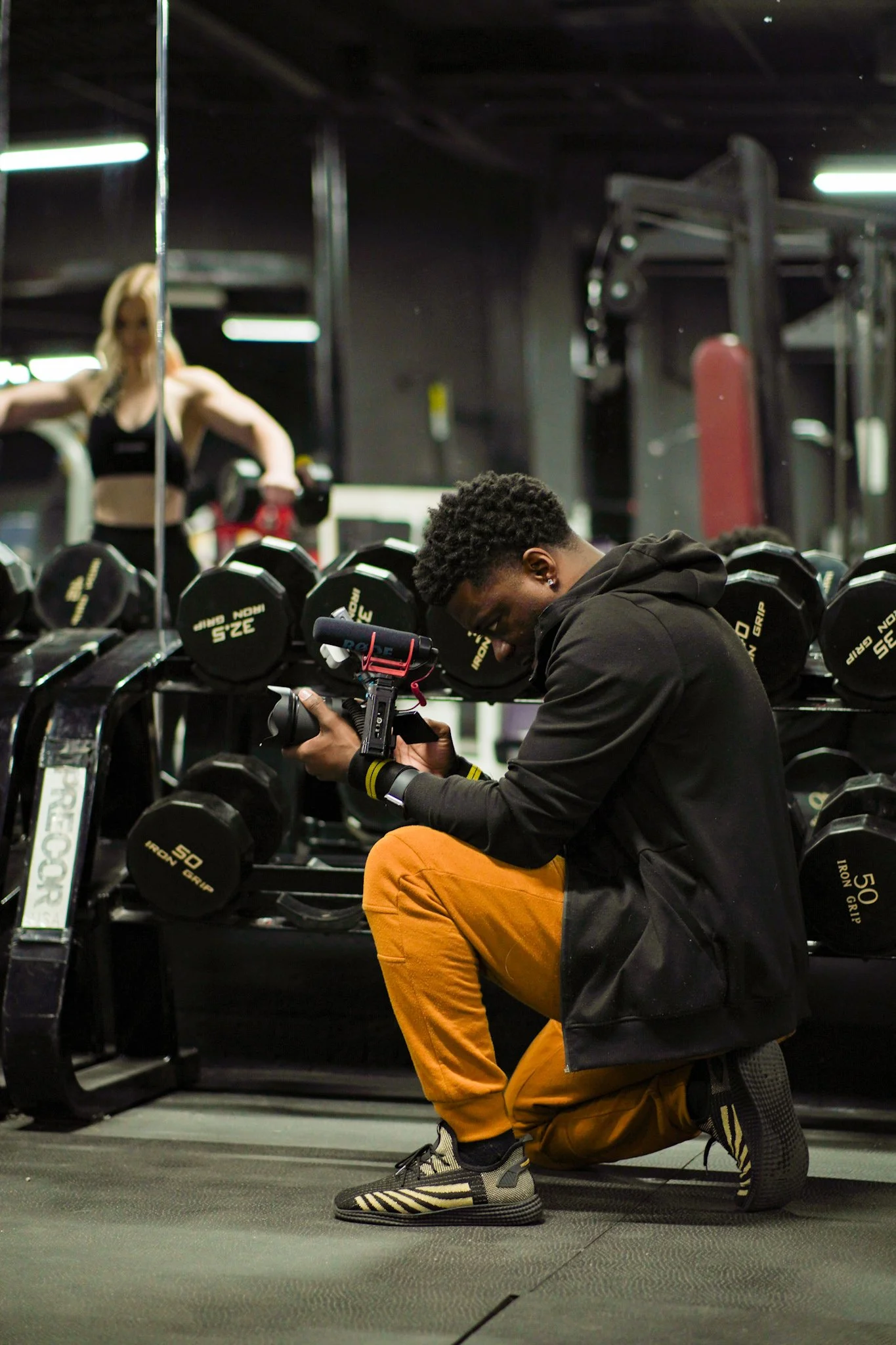 A man kneeling on the gym floor, holding a camera, with a row of dumbbells behind him and a woman lifting weights in the background.