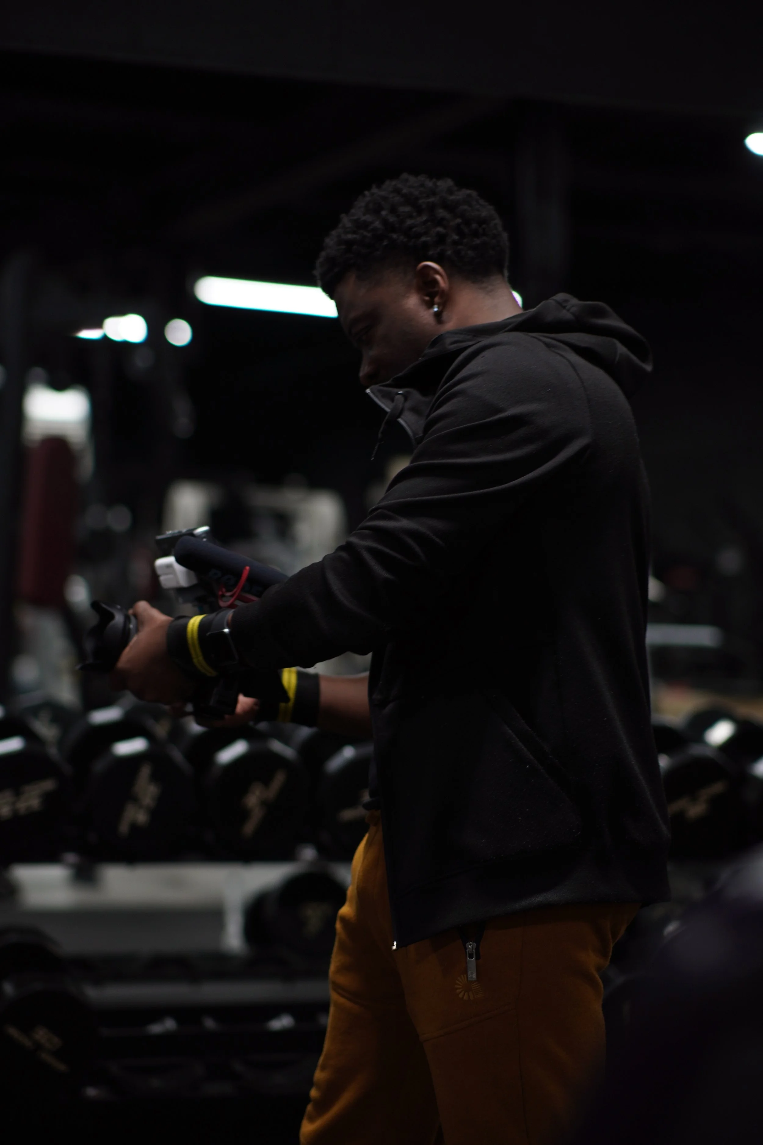 A young man in a black hoodie and brown pants standing in a dimly lit gym, looking at his phone with weightlifting equipment in the background.