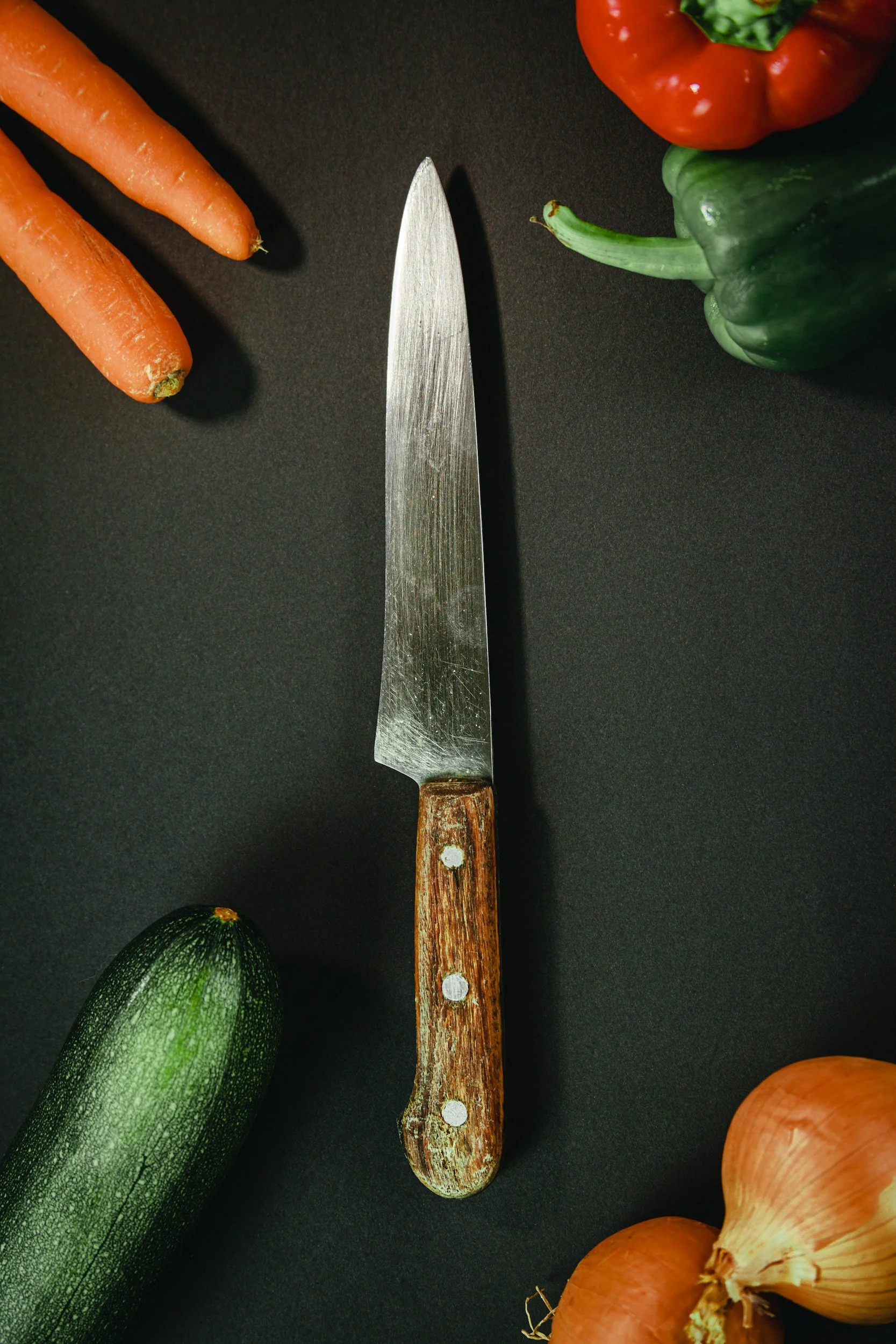 A kitchen knife with a wooden handle is placed on a black surface surrounded by fresh vegetables, including carrots, a red bell pepper, a cucumber, an onion, and a green bell pepper.