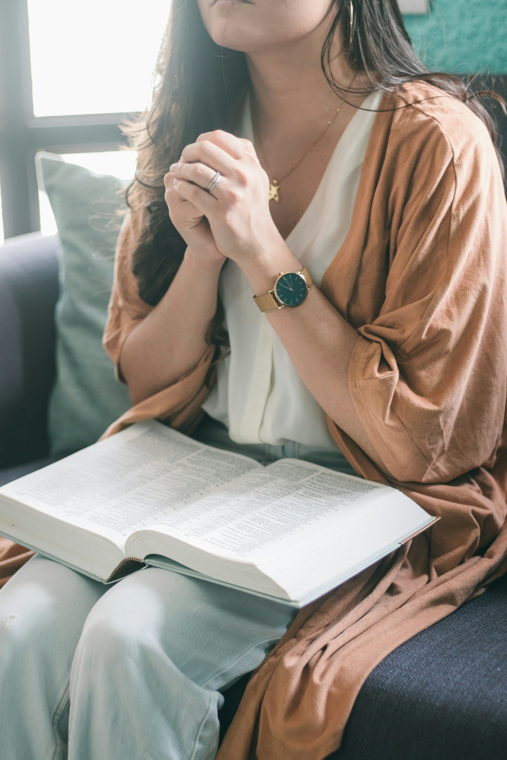 A woman with long dark hair wearing a white dress, a gold watch, a ring, and a gold necklace, sitting on a couch with a large book open on her lap, clasping her hands together in prayer.