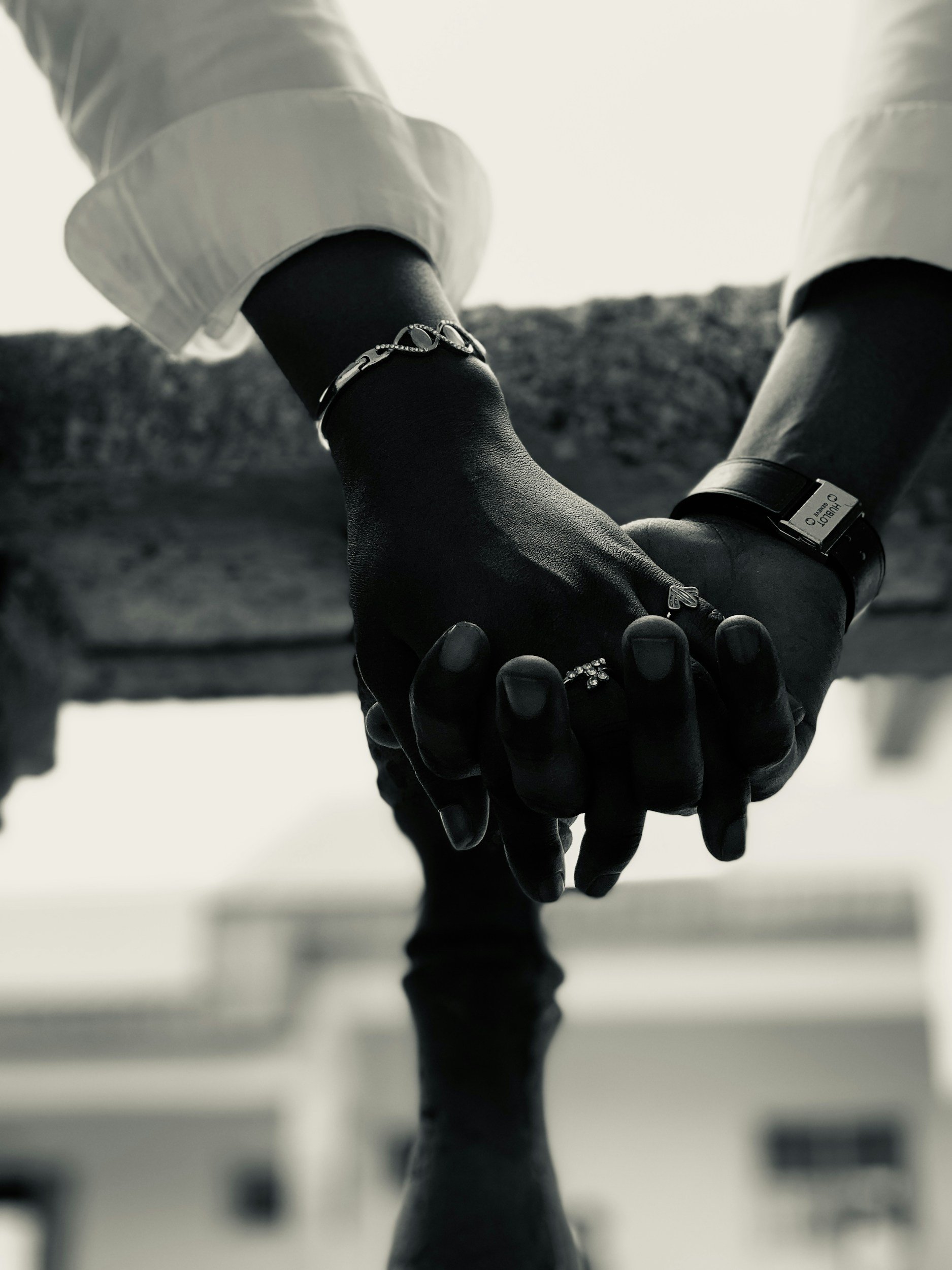 Close-up of two people holding hands, one wearing a black leather glove with rings and a bracelet, and a white shirt sleeve, against a bright sky background.