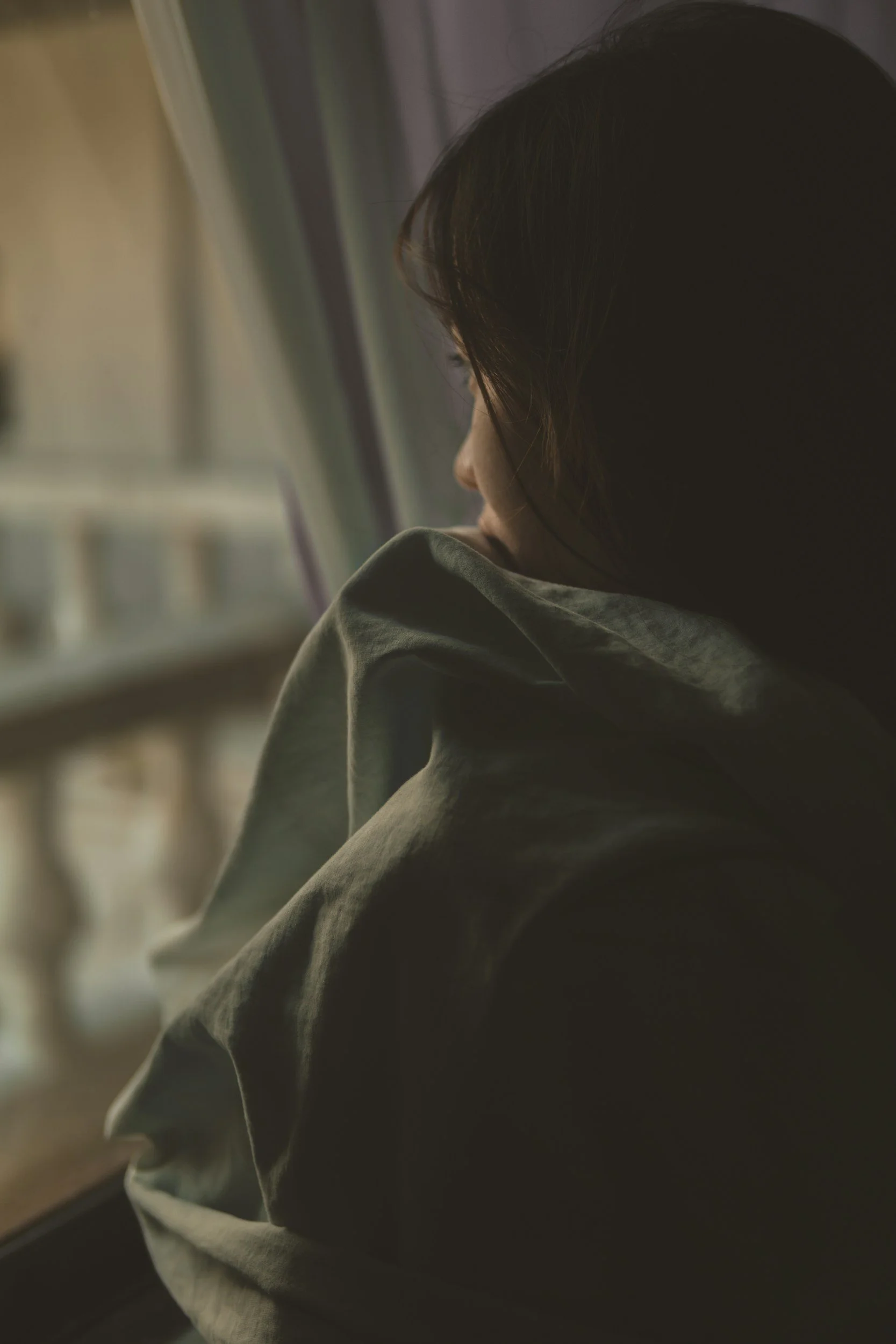 A person with dark hair sitting with their head tilted towards a window, wearing a gray hoodie, in a dimly lit room.