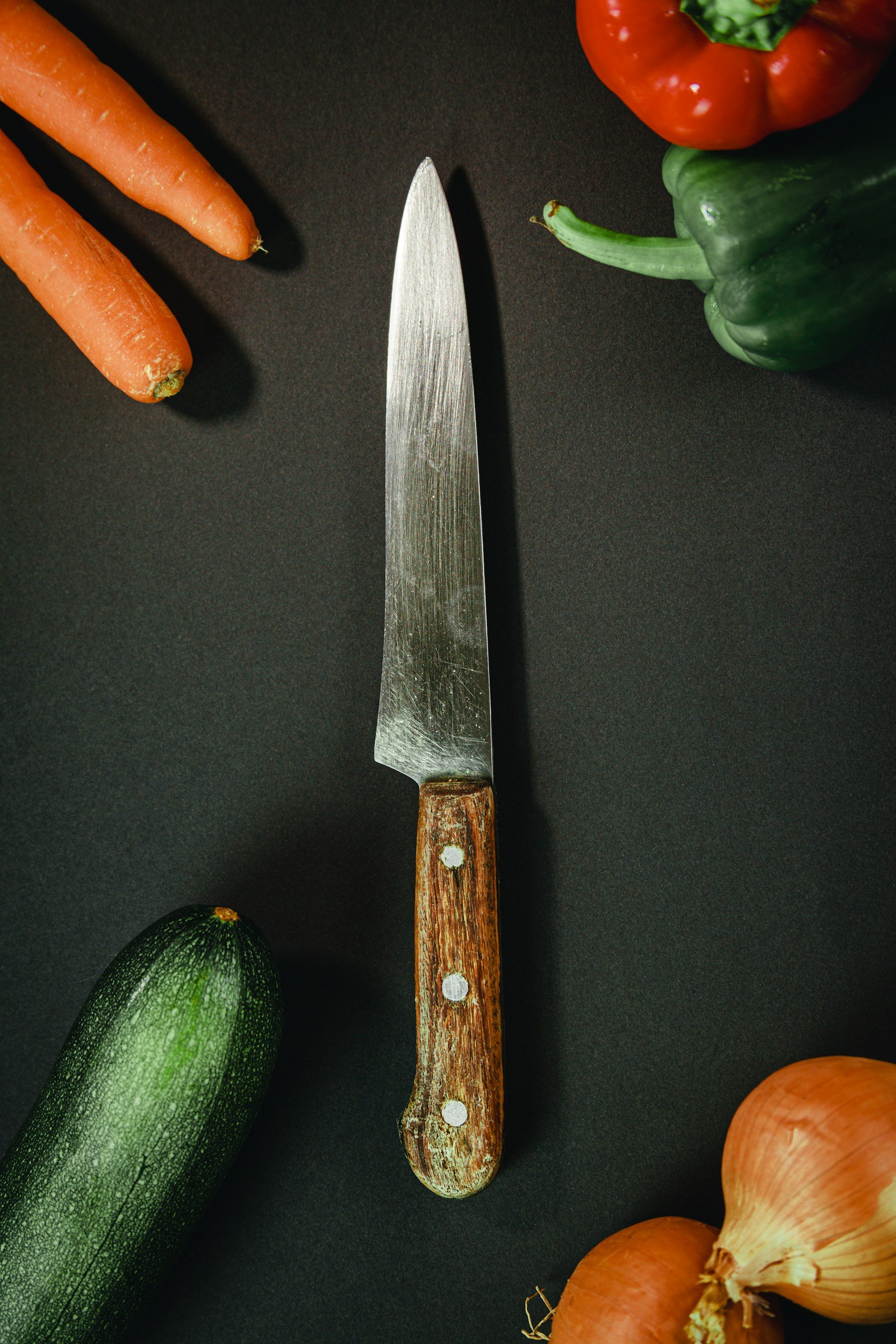 A kitchen knife with a wooden handle centered between a zucchini, carrots, onions, and bell peppers on a dark surface.