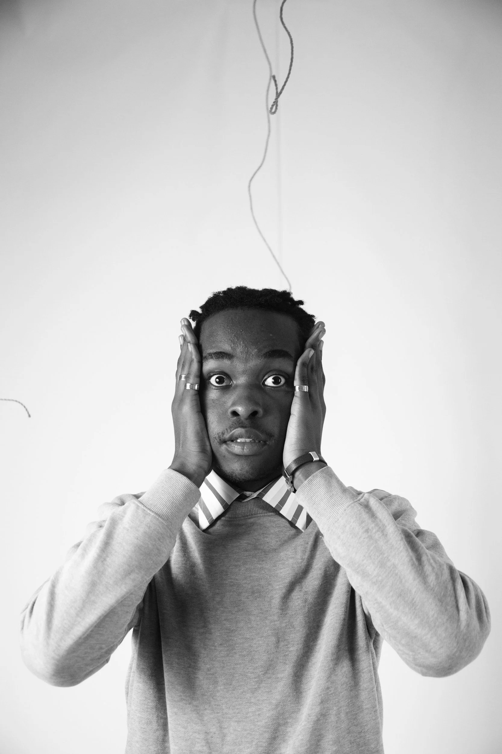 A young Black man with short curly hair and expressive eyes holds his head with both hands, looking directly at the camera. He is wearing a light-colored sweater over a collared shirt with striped pattern. The background is plain and white, and there is a thin wire hanging from the top of the image.