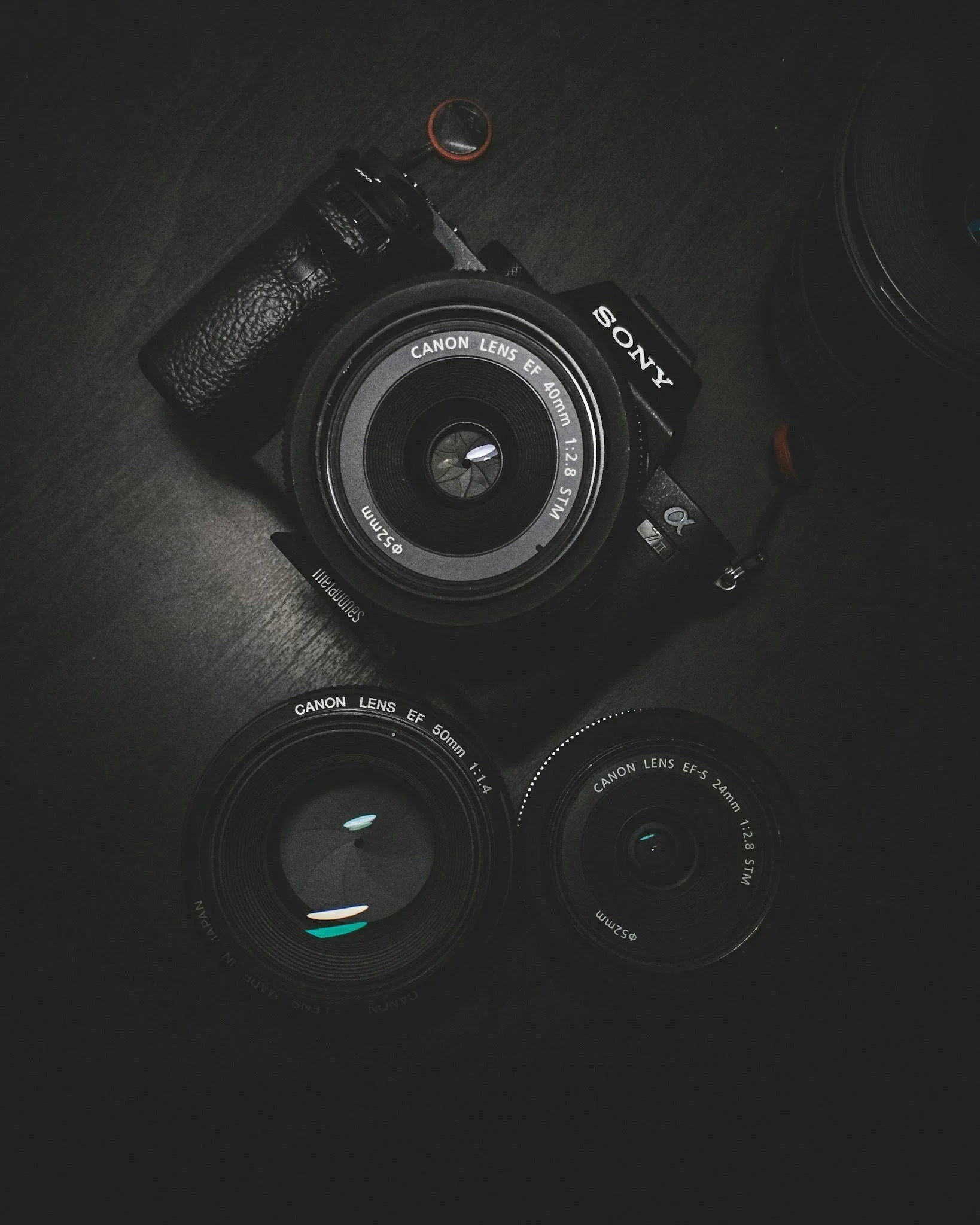 Photograph of a Canon camera with 40mm lens on a dark surface, accompanied by multiple camera lenses and lens caps.