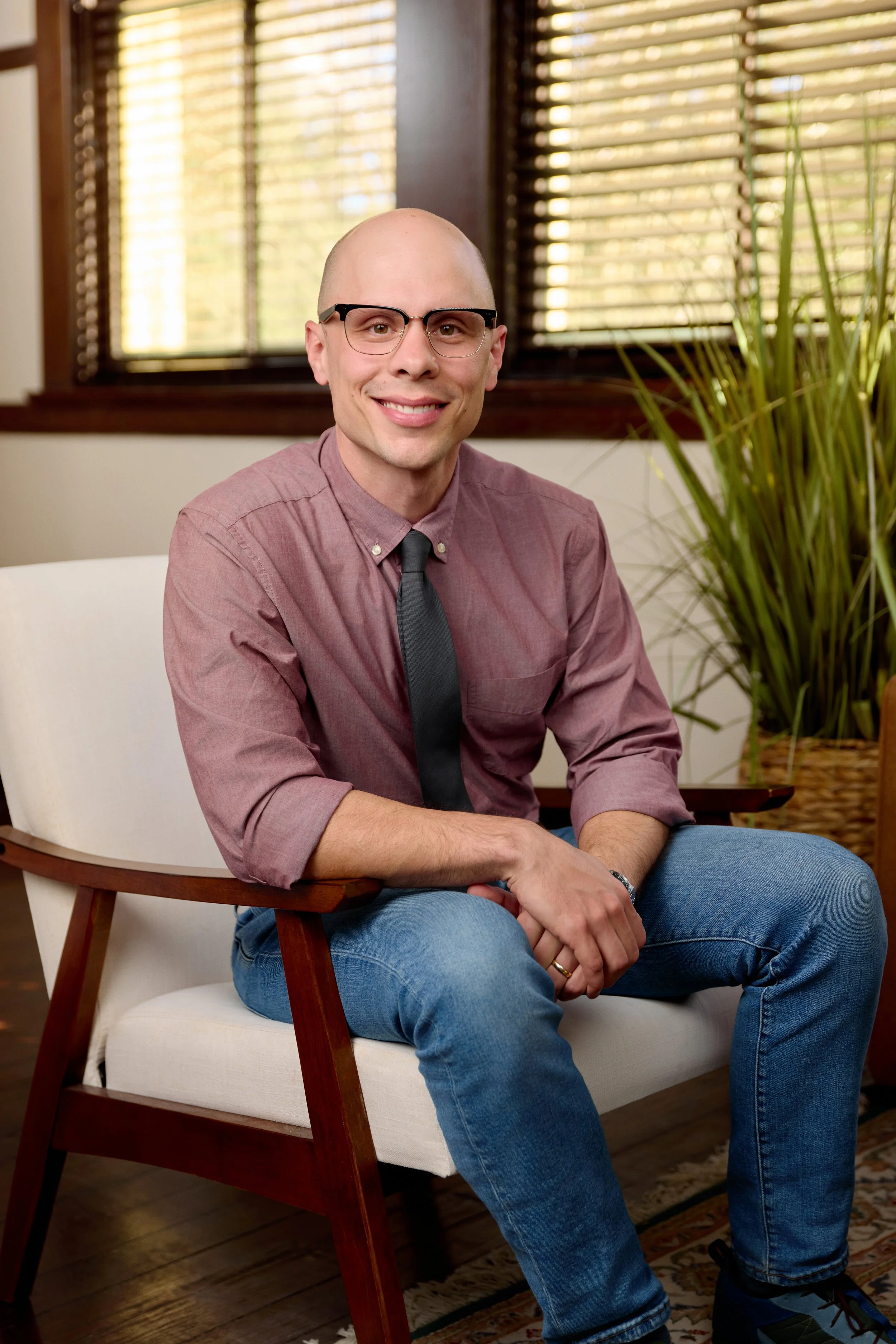 A man with a shaved head and glasses sitting on a white chair in front of a window with wooden blinds, wearing a mauve button-up shirt, black tie, and blue jeans, smiling at the camera. There is a large plant visible on the right side of the image.