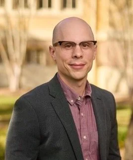 A man wearing glasses and a dark blazer standing outdoors with trees and a building in the background.