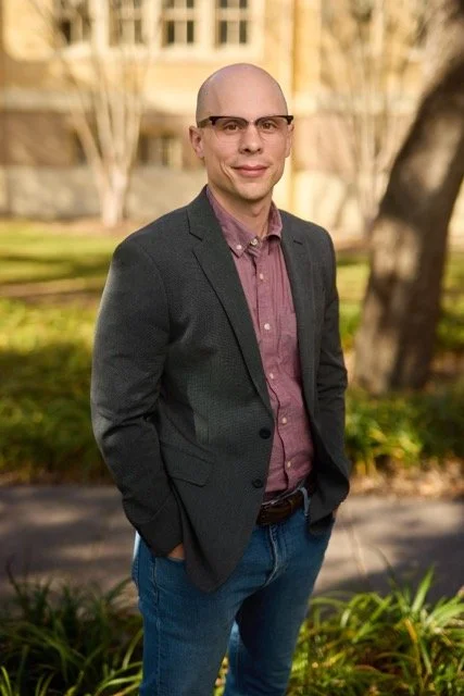 A man with glasses, a bald head, wearing a dark blazer and maroon shirt, standing outdoors in a park with trees and a building in the background.