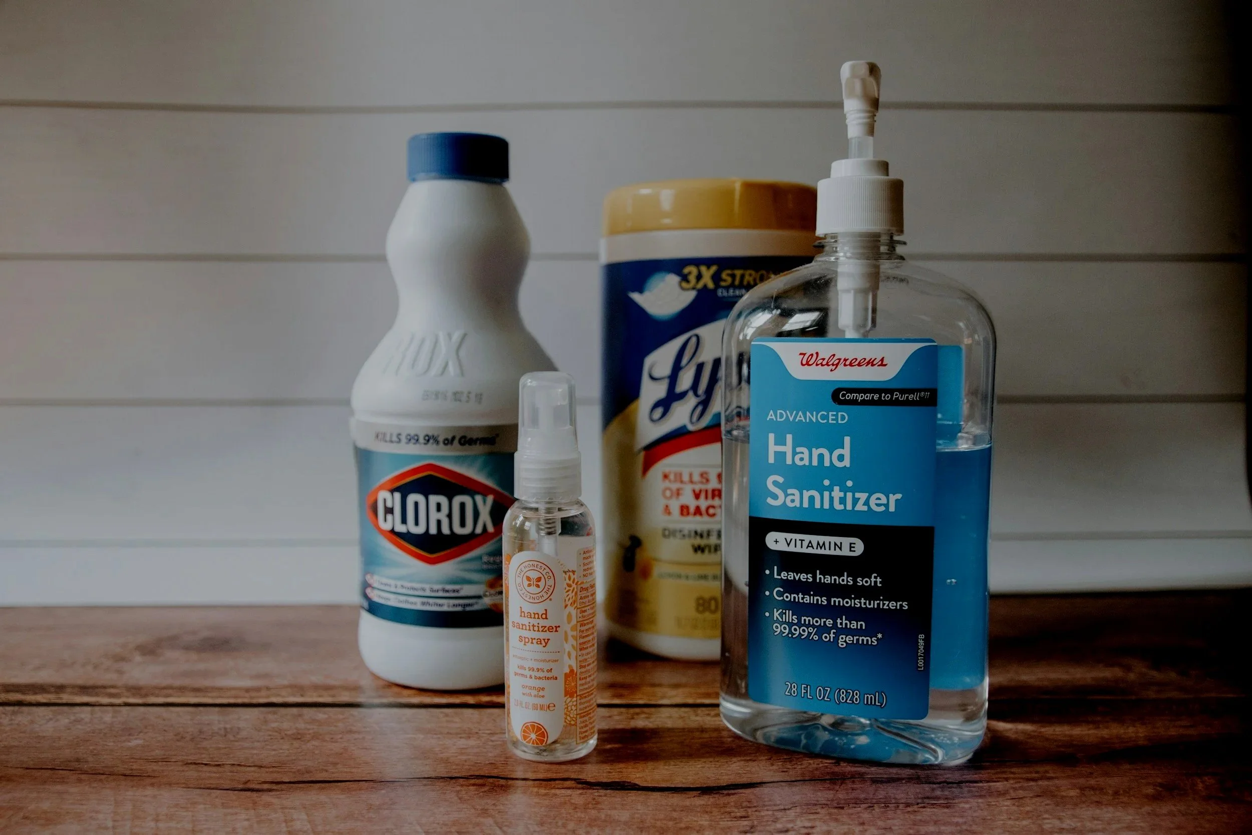 Various hygiene and cleaning products on a wooden surface, including hand sanitizer, disinfectant spray, and wipes, against a white shiplap background.