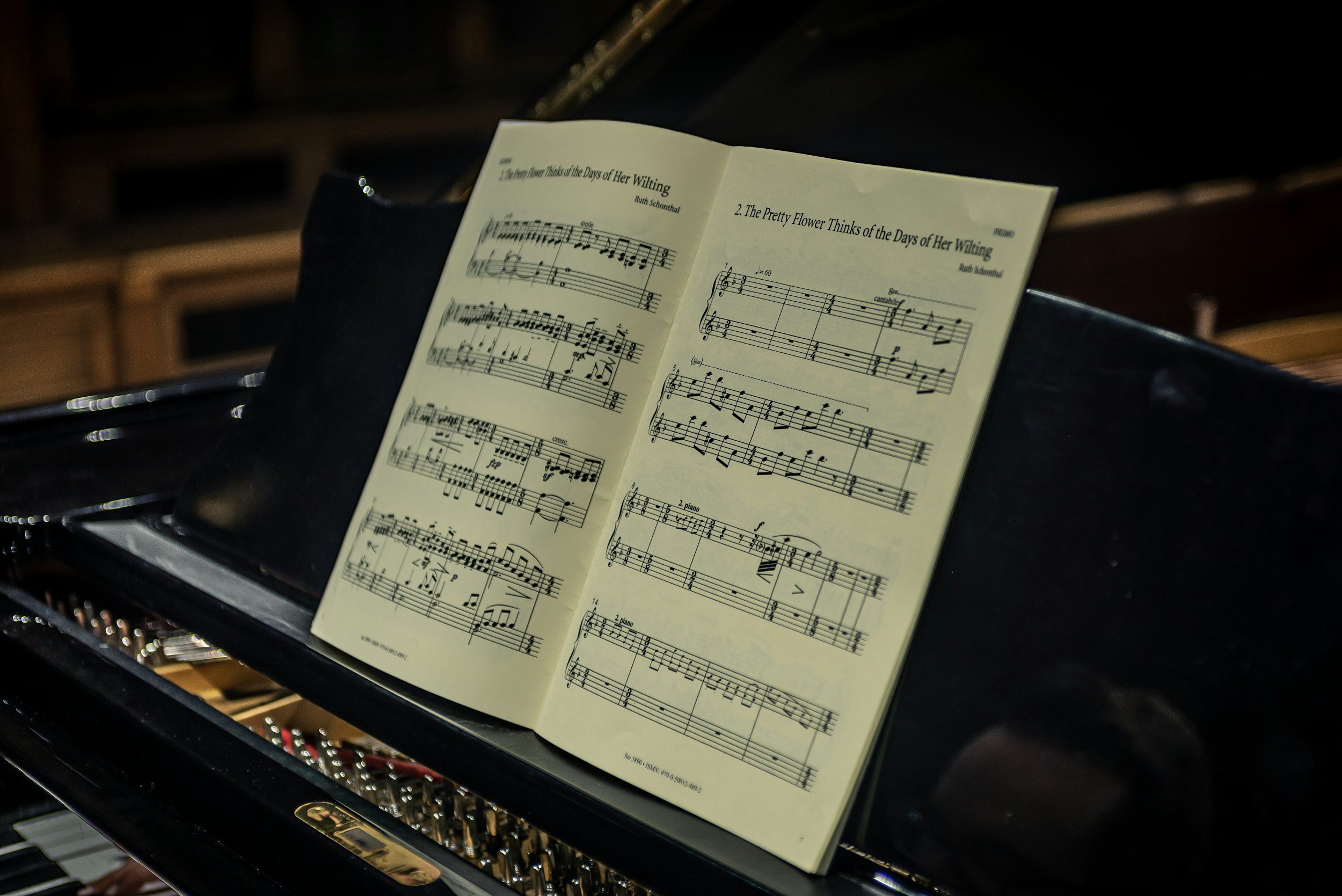Sheet music for a piano piece titled 'The Pretty Flower Thinks of the Days of Her Waiting' by Ruth Schoenthal, resting on a grand piano with visible interior strings.