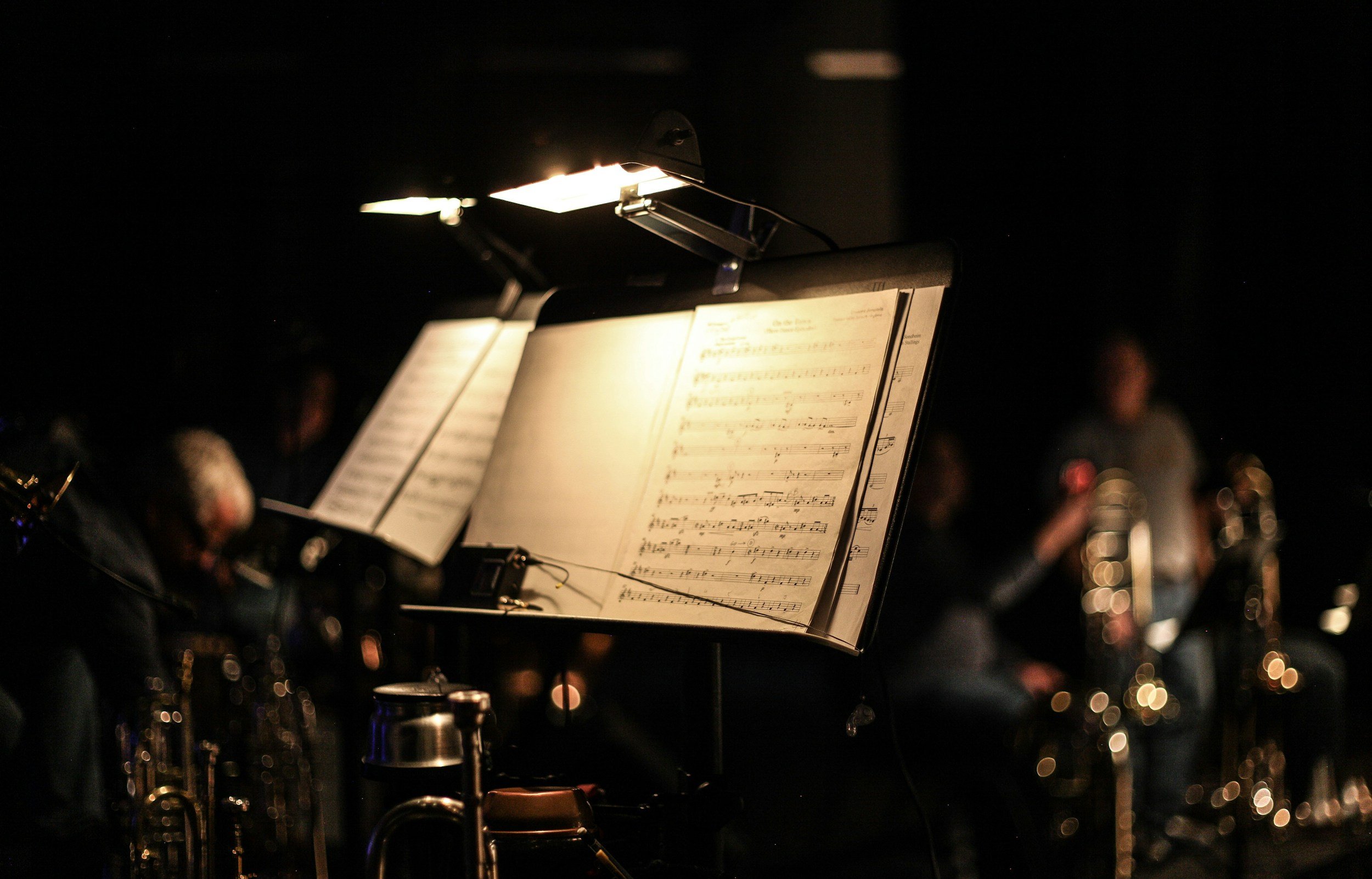 A music stand with sheet music illuminated by a small light, with blurred musicians and instruments in a dark setting.