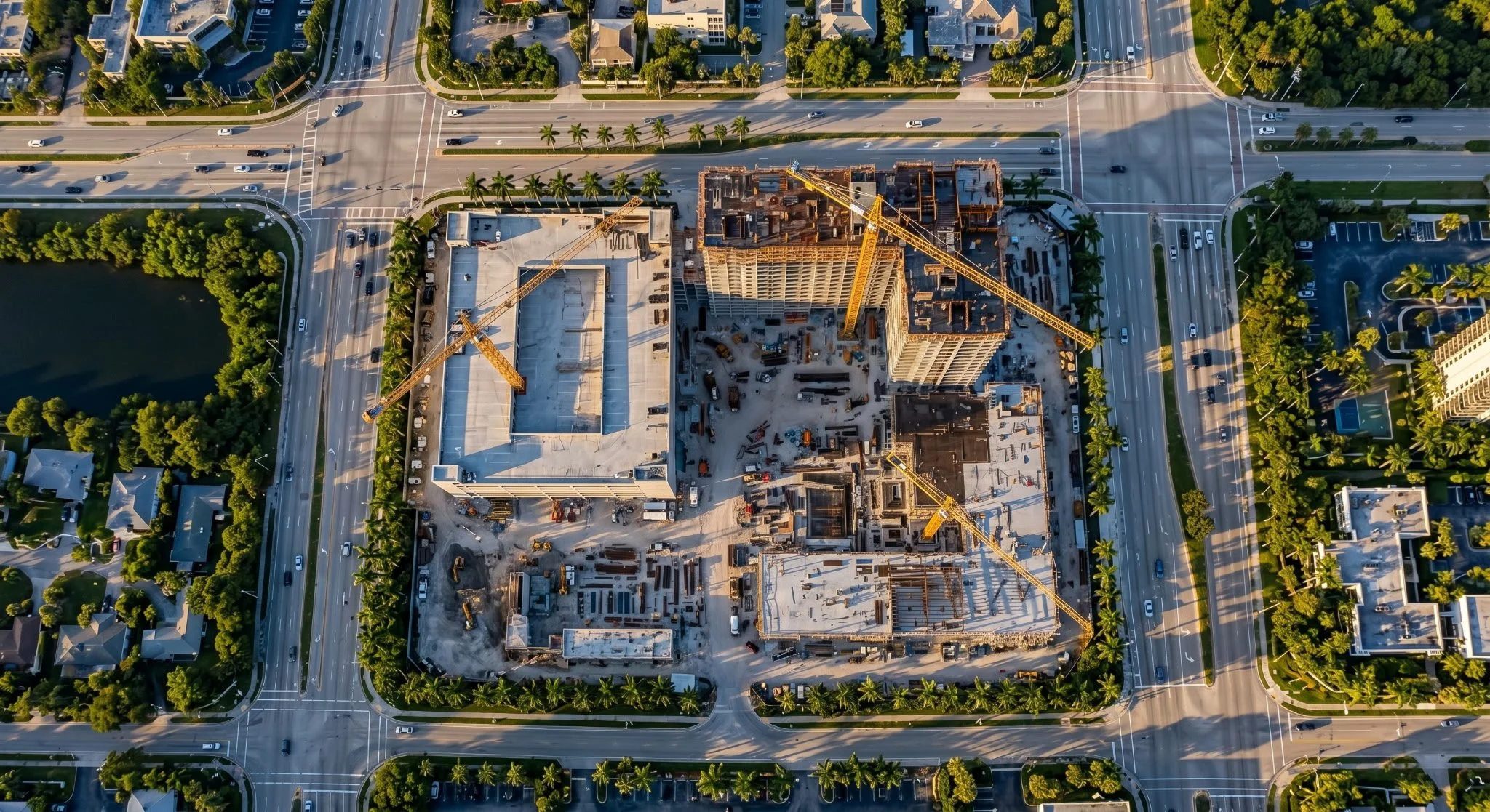 An aerial view of a construction site in a city with cranes and partially built high-rise buildings, surrounded by roads with cars and nearby residential houses and trees.