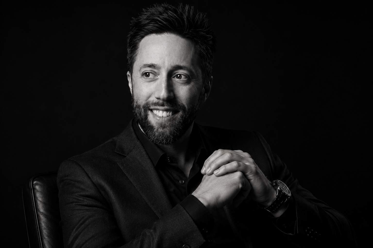 Black and white portrait of a man with a beard, smiling, with his hands clasped, wearing a dark blazer and a wristwatch, seated on a chair against a dark background.