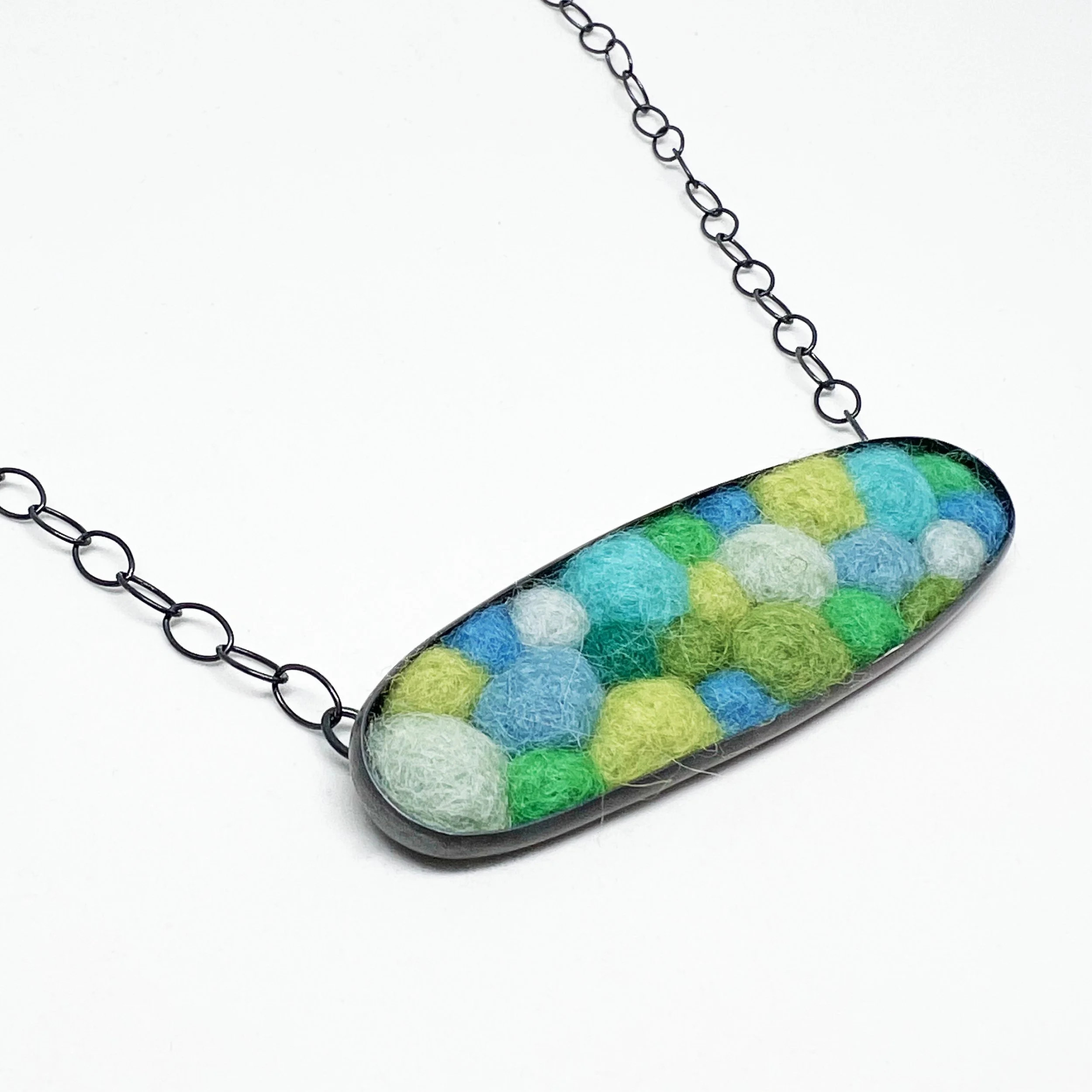Close-up of a colorful, oval-shaped pendant with a pattern of blue, green, and yellow blotches, attached to a black metal chain, against a plain white background.