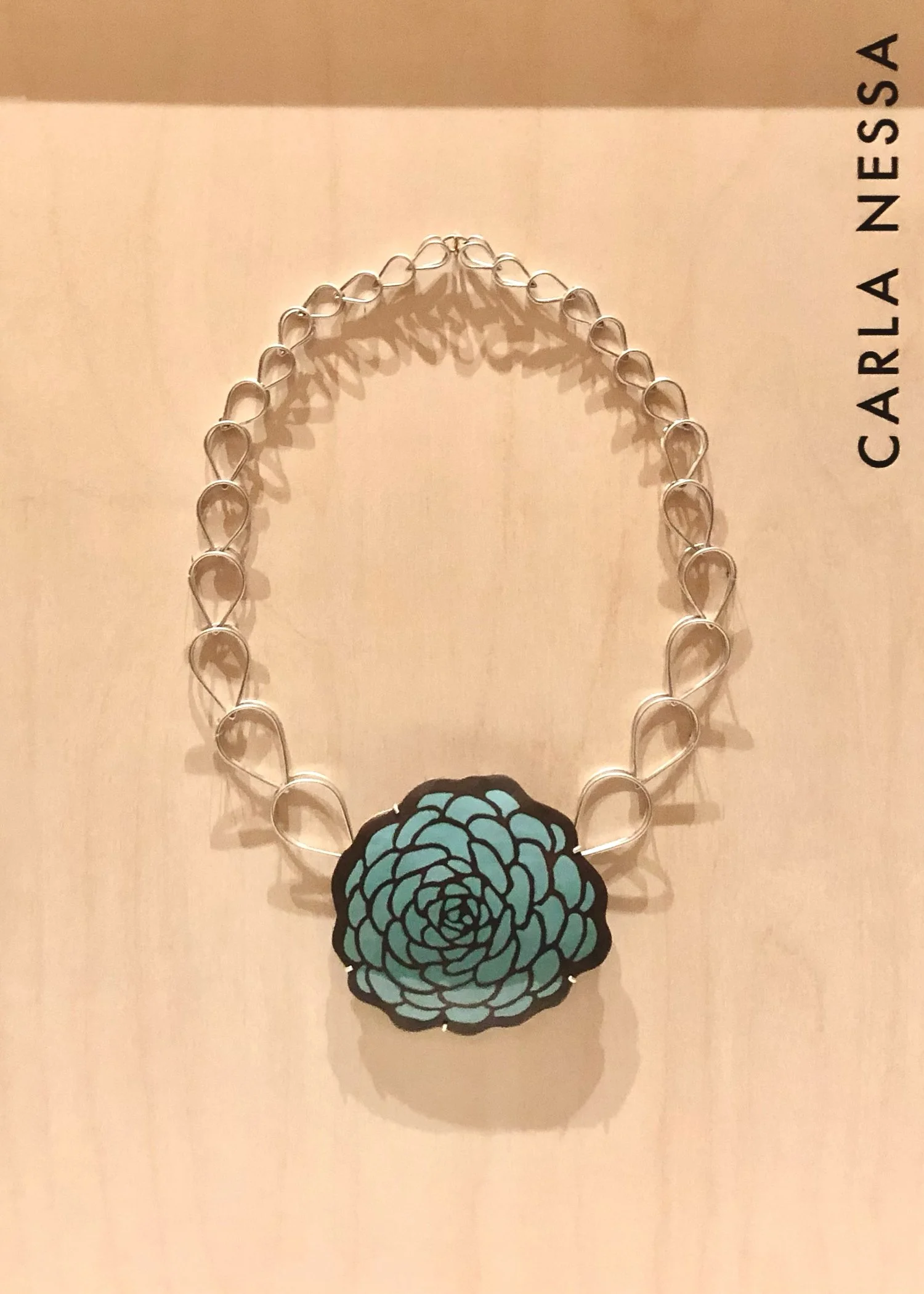 A necklace with large, open, metallic heart-shaped links, a flower-shaped pendant with black outlines, and a turquoise floral design. The necklace hangs against a light beige background.