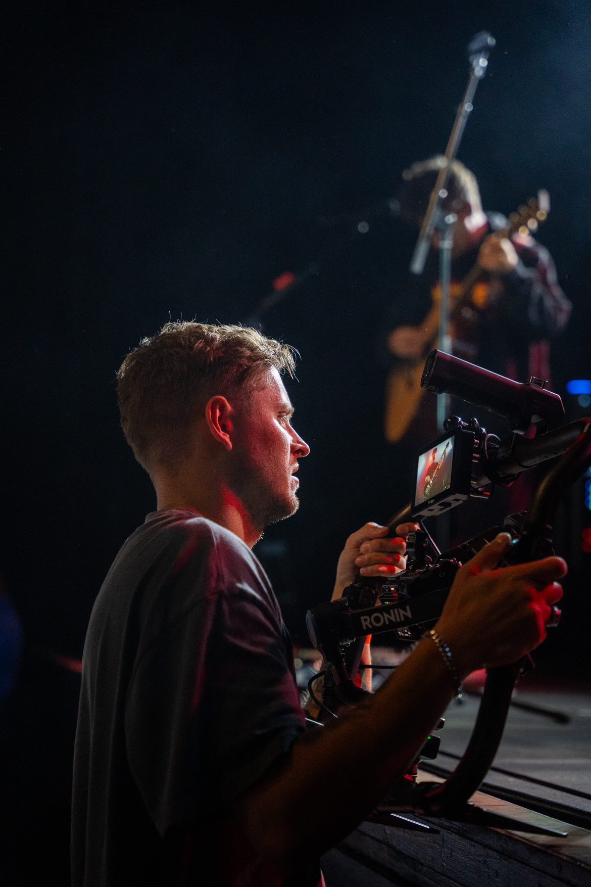 Ein Mann filmt mit einer Kamera, im Hintergrund ein Musiker mit Gitarre bei einem Auftritt während der Nacht.