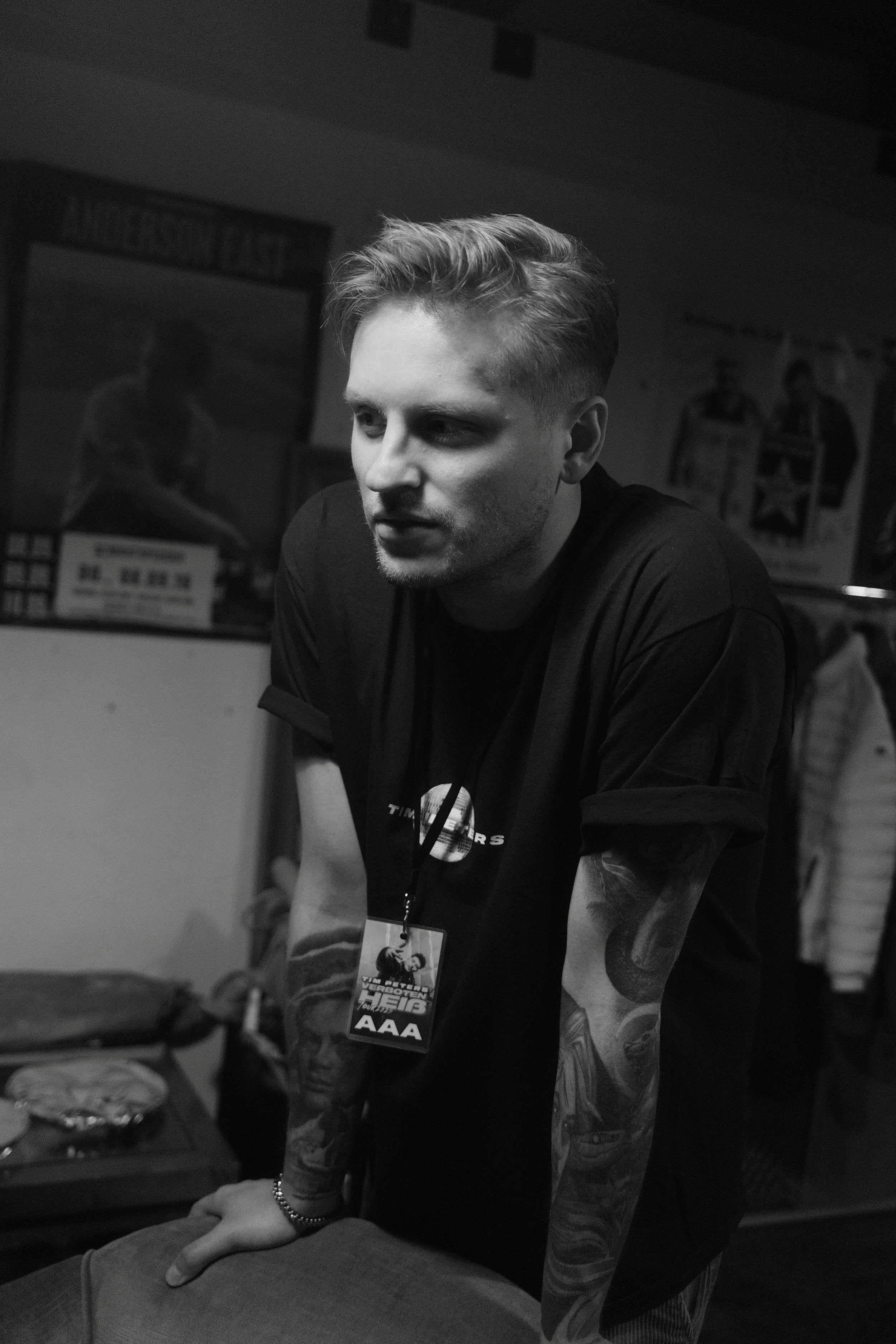 Schwarz-weißFoto eines jungen Mannes mit kurzen, stylischen Haaren und Tattoos, der eine Karte trägt, auf denen 'AAA' steht, in einer Indoor-Umgebung mit Postern im Hintergrund.