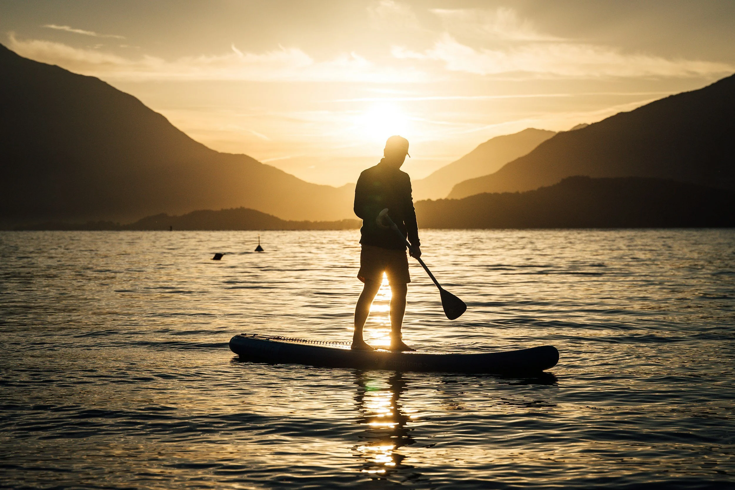 Person paddleboarding at sunset on a calm lake with mountains in the background.