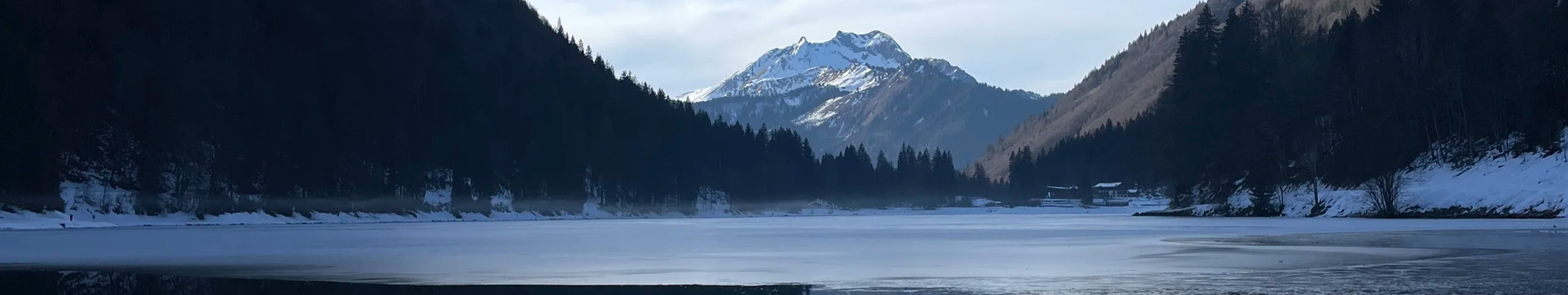 Snow-covered mountains and dense evergreen trees surrounding a partially frozen alpine lake.