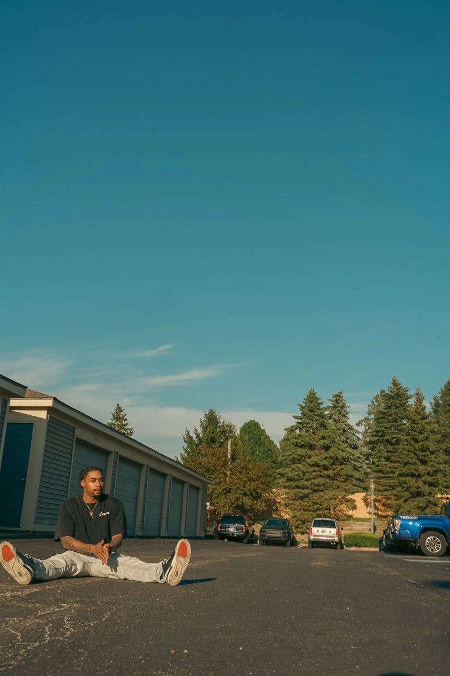 A young man sitting on the pavement in a parking lot, with a row of parked cars and trees behind him under a clear blue sky.