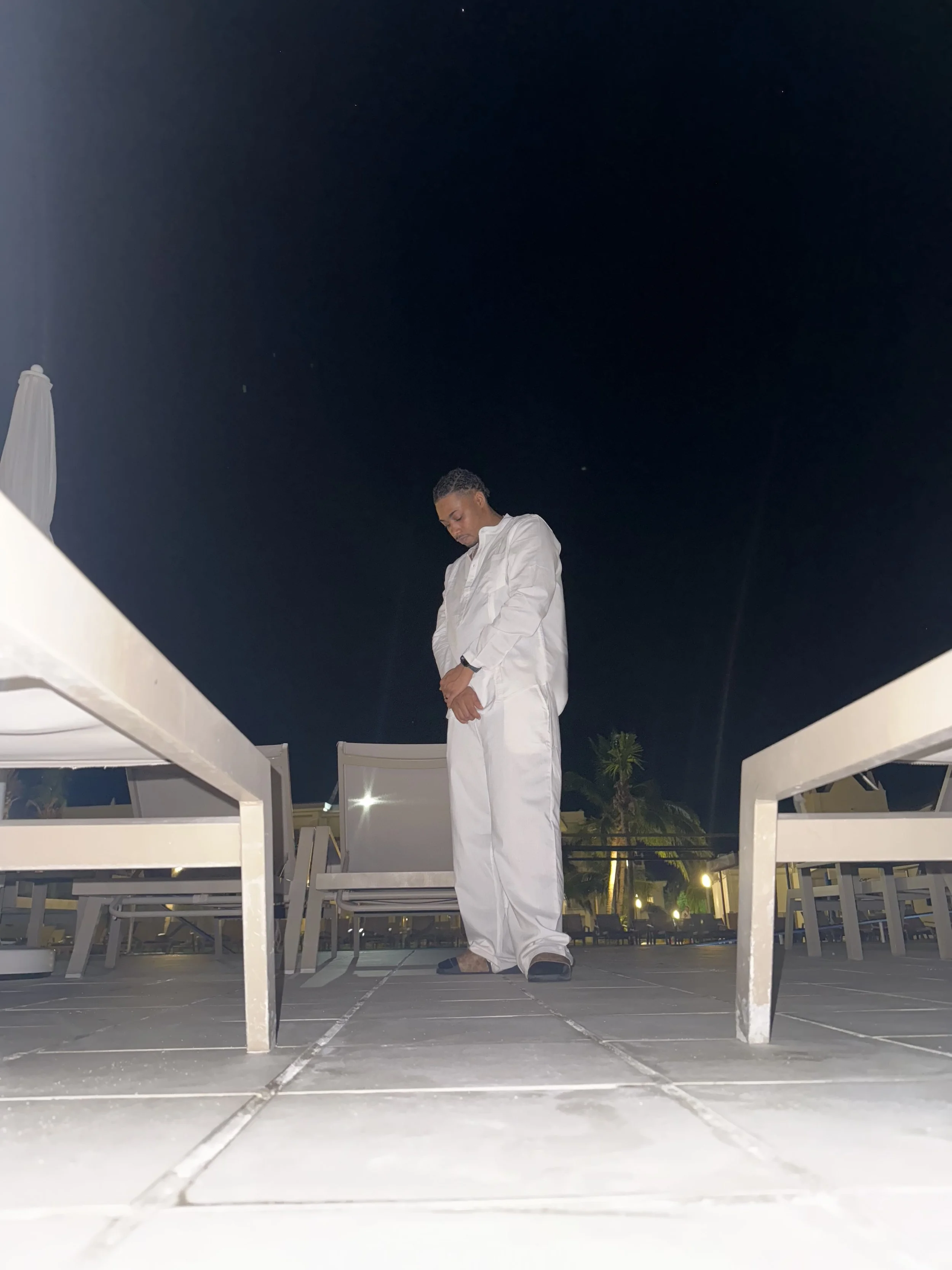 A man dressed in white standing outdoors at night, looking down with a serious expression. The scene includes white outdoor furniture and a dark sky with some visible stars.