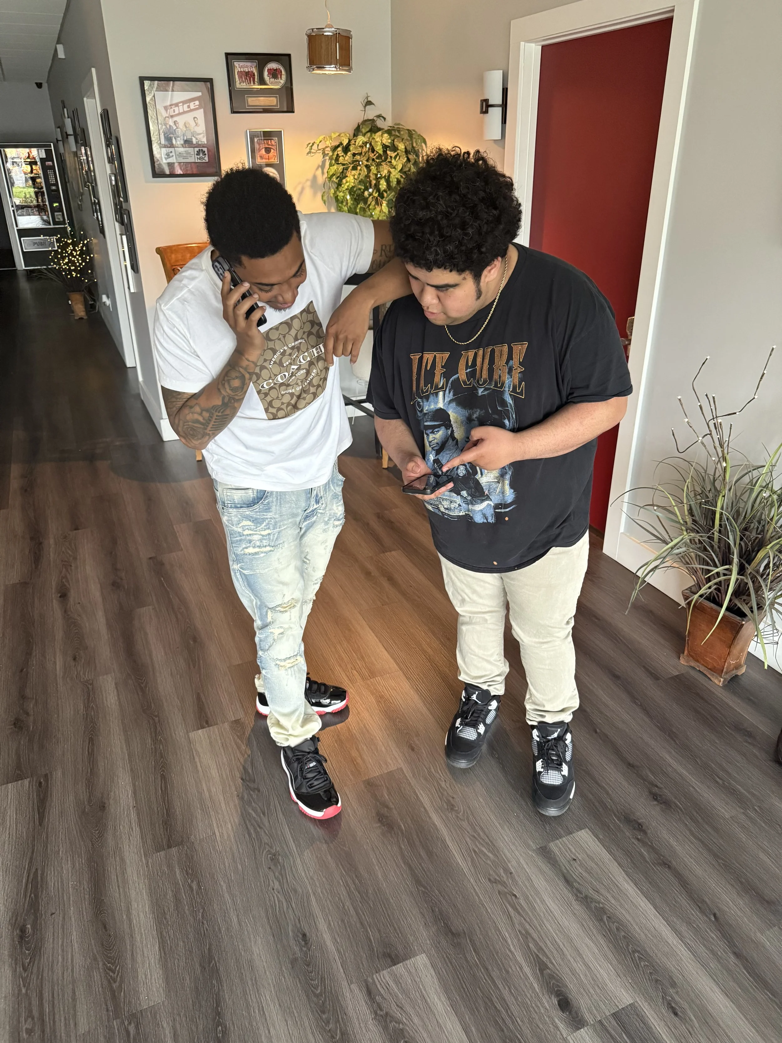 Two young men standing on a wooden floor, examining something on a smartphone held by the man on the right, while the man on the left talks on a landline phone. They are inside a house with framed pictures on the wall, a decorative plant, and a plant